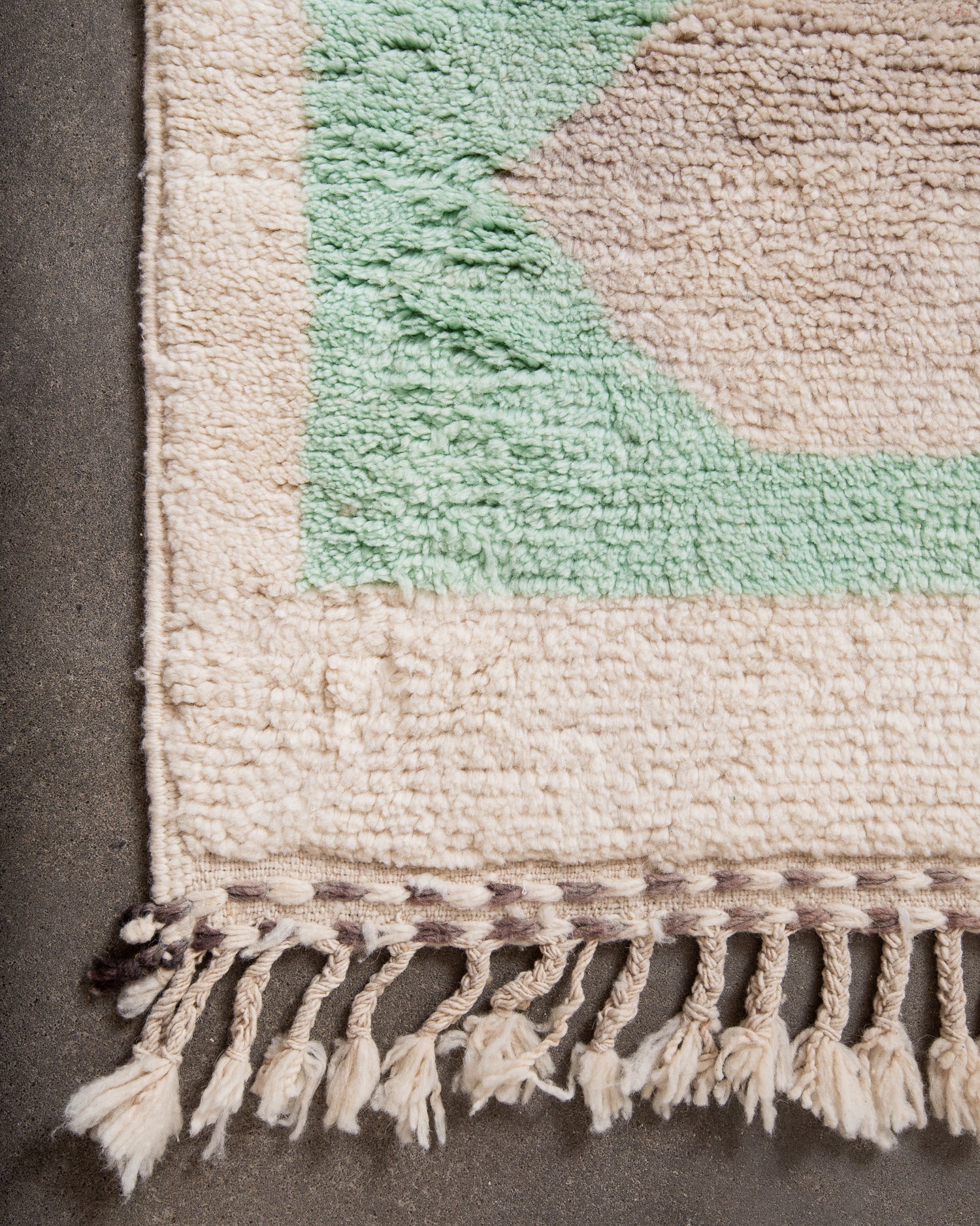 Corner of a textured rug with tassels on a cement floor.