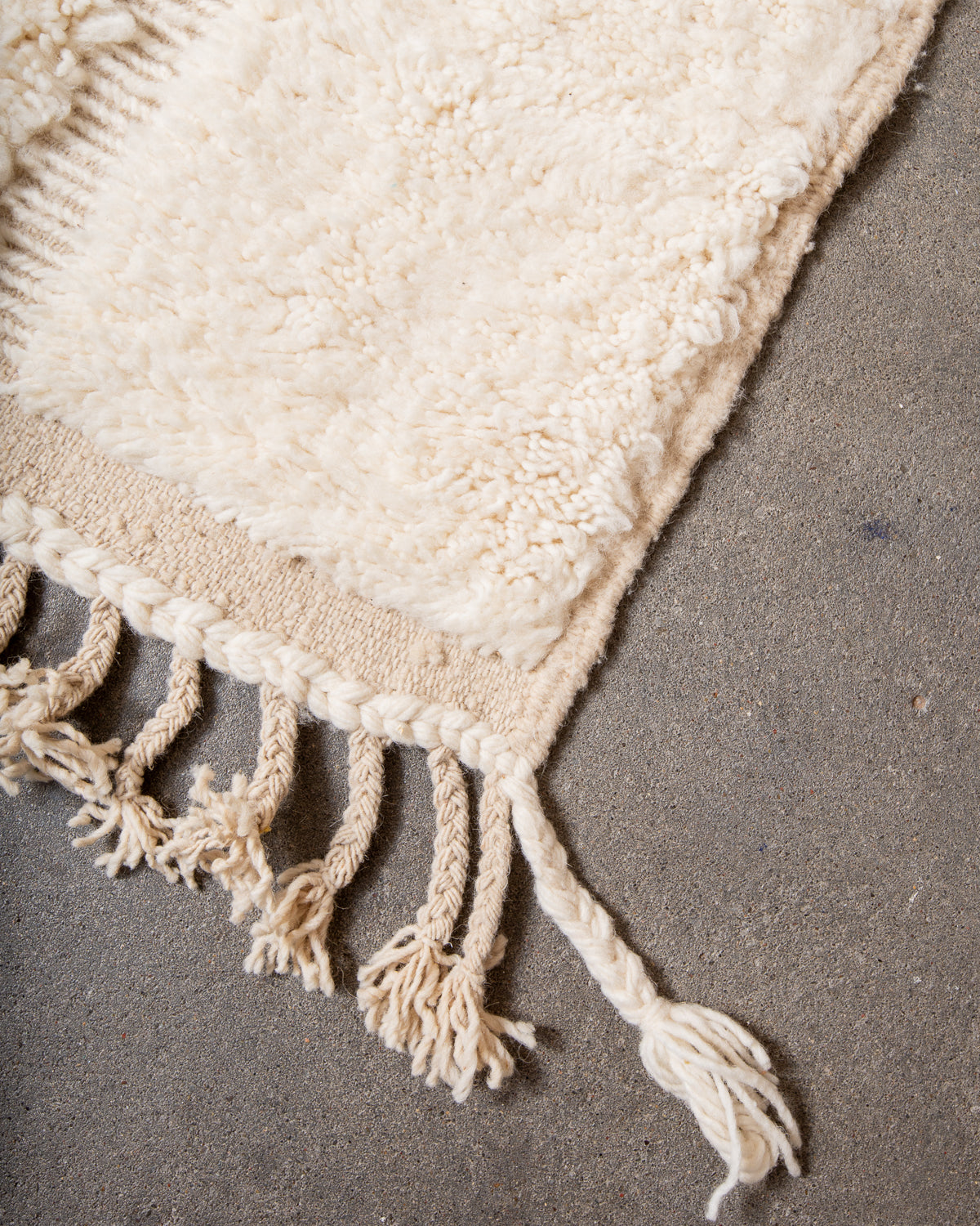 Corner of cream wool rug with tassels on grey cement floor. 
