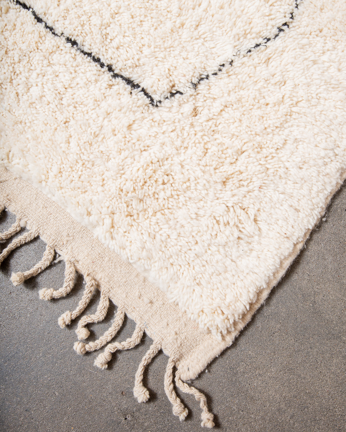 Corner of cream wool rug with tightly woven end and natural wool tassels on grey floor.