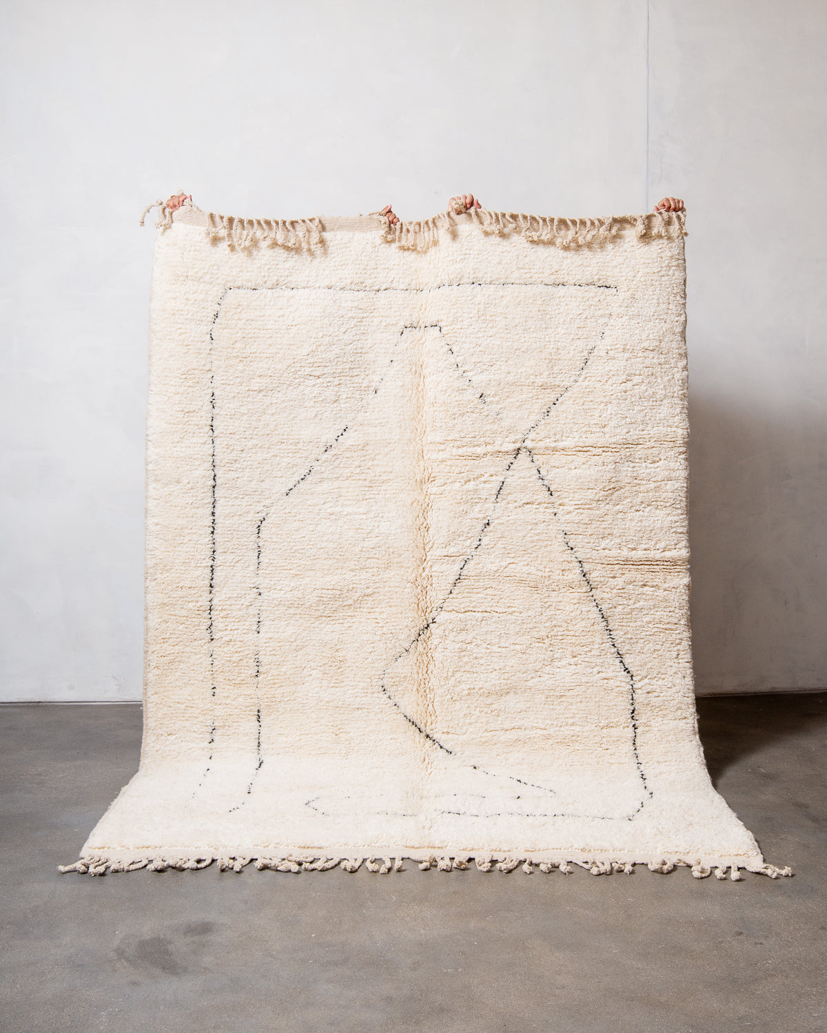 Cream wool rug with black intersecting lines being held up in front of a white wall.