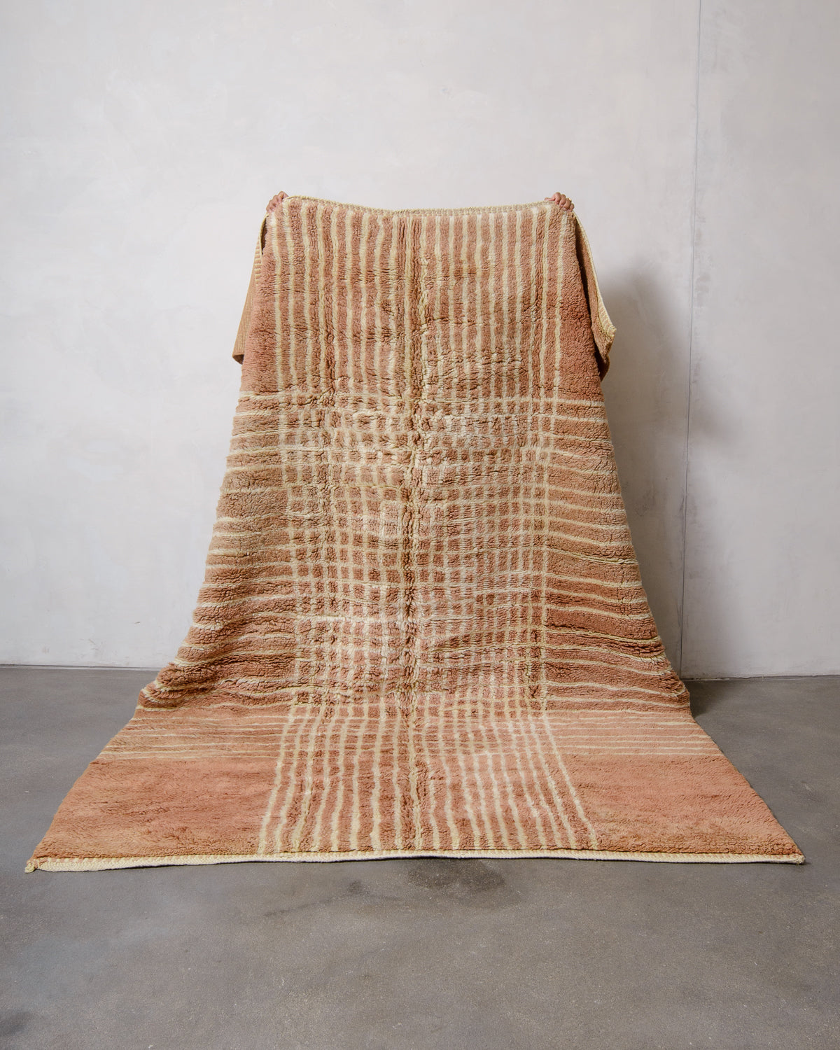 Berberlin Beni M'rirt rug in camel brown with lemon cream coloured grid pattern held up by two hands against white wall and grey floor.