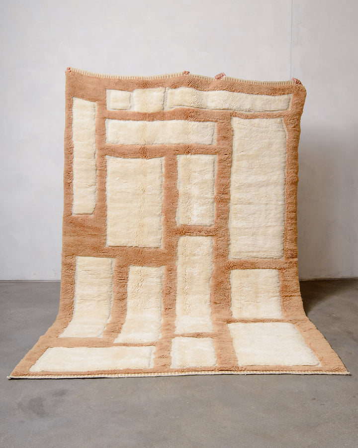 Berberlin Beni M'rirt wool rug in cream and light brown geometric design, being held up against white wall and grey cement floor.