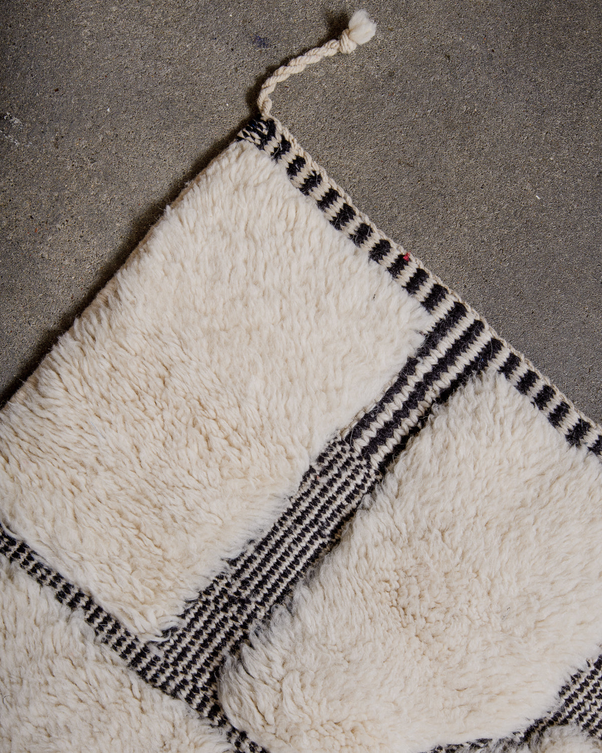 Corner of cream M'rirt rug with tightly woven edges with black and white striped pattern and one corner tassel.