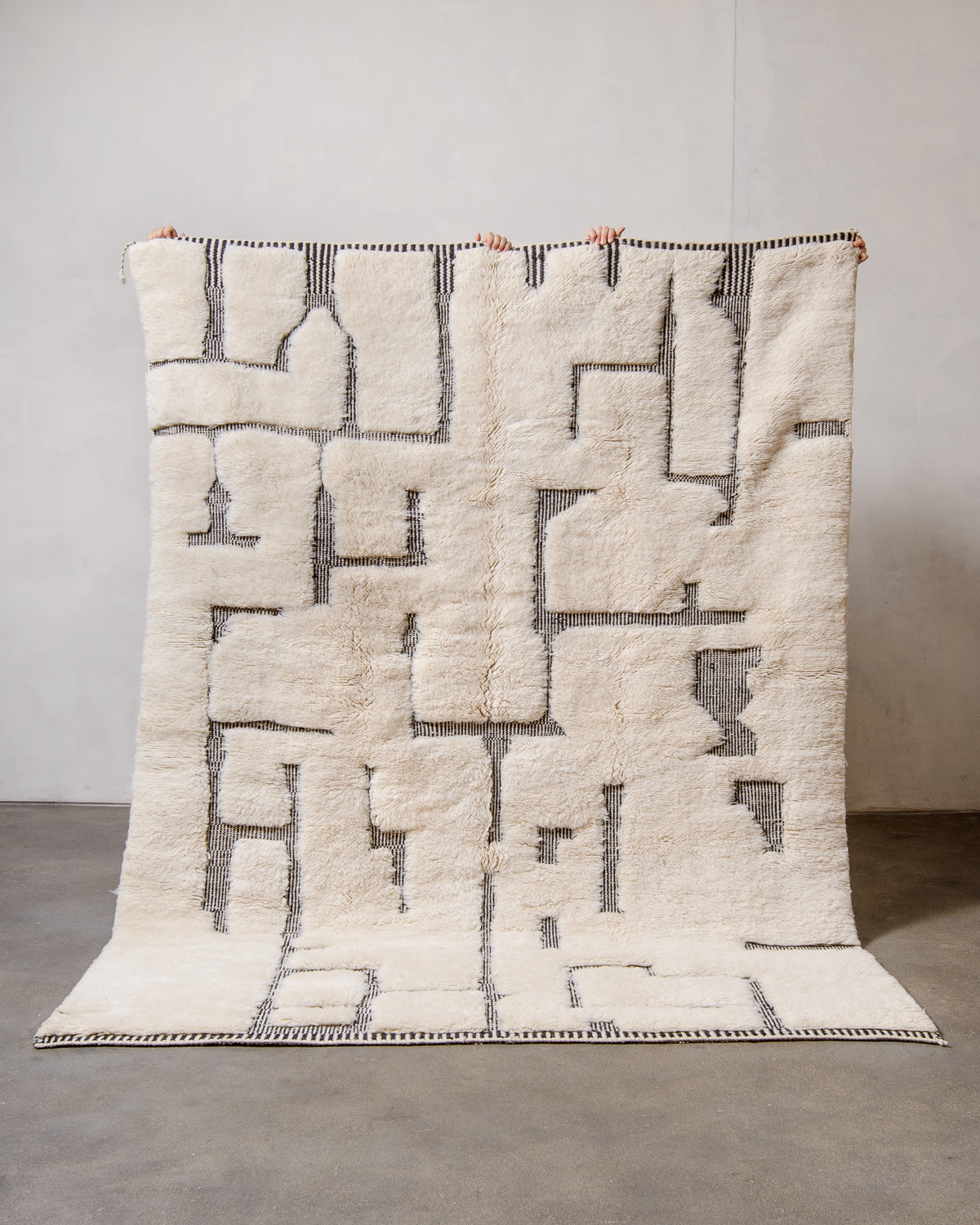 Cream M'rirt Rug with abstract pattern in woven black stripes being held in front of a white wall. 