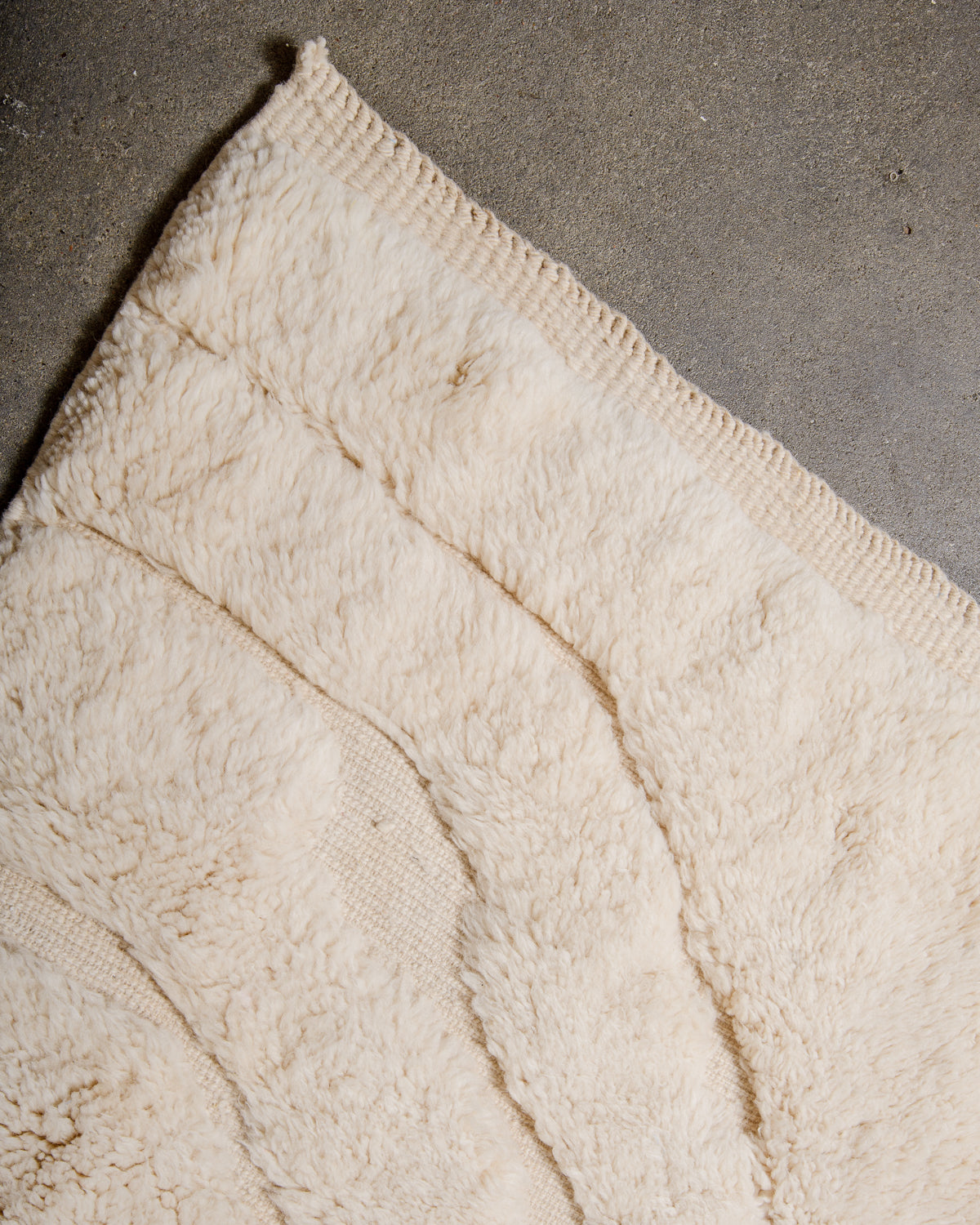 Corner of Beni M'rirt rug in cream wool showing tightly woven end laying on concrete floor.