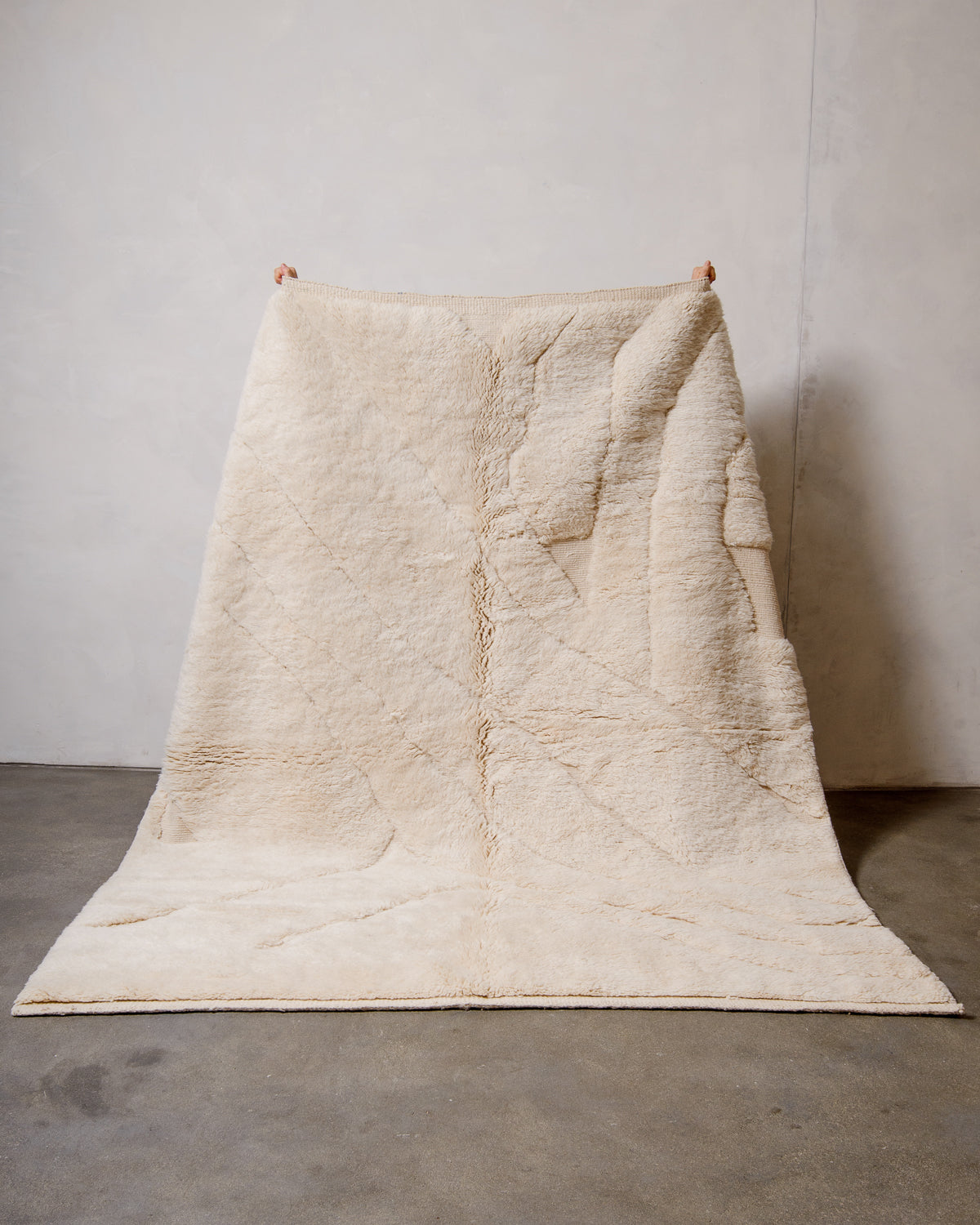 Berberlin Beni M'rirt rug in cream wool with abstract woven design, being held up against a white wall in a studio.