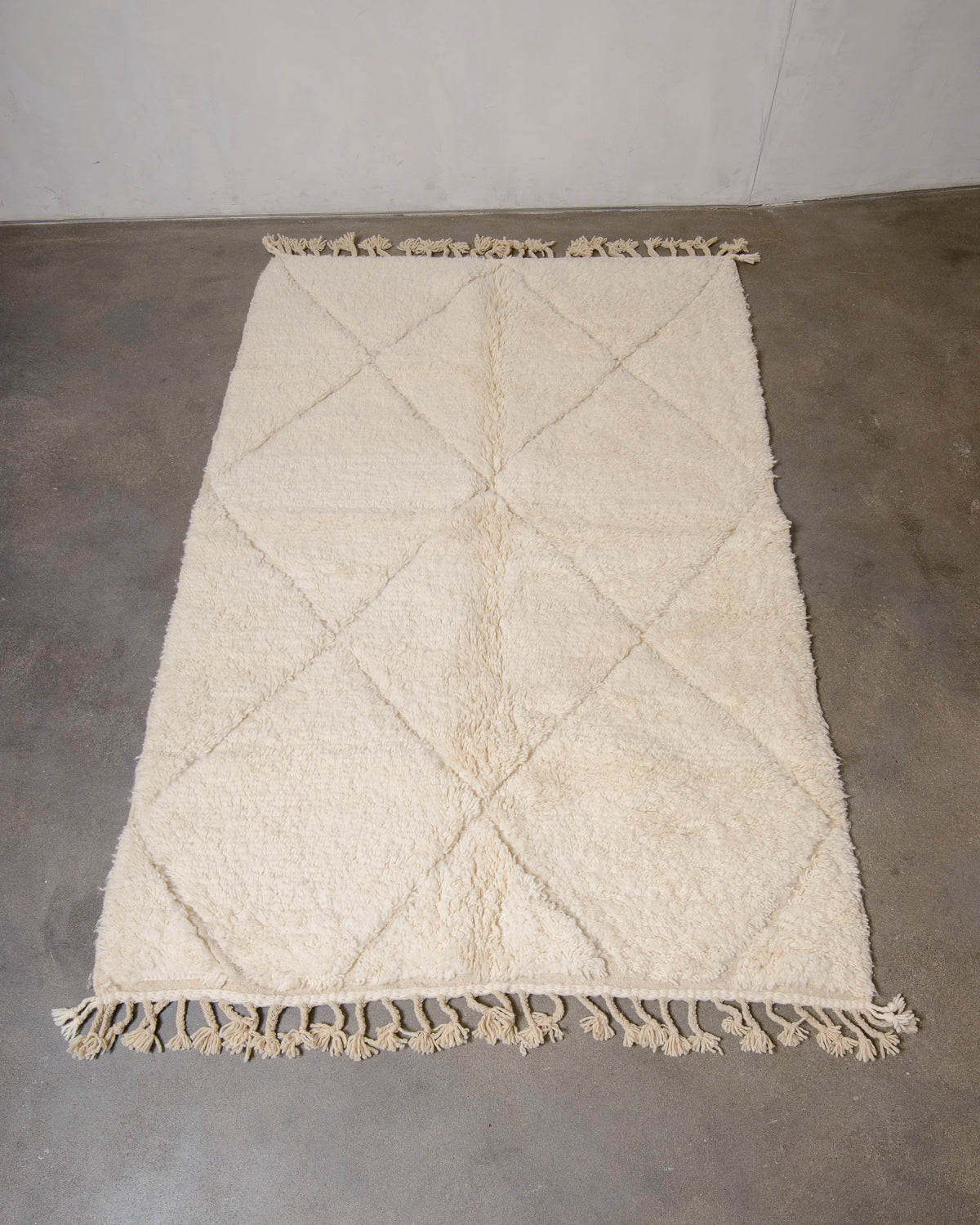 Cream wool rug with woven diamond pattern and natural wool tassels at each end, on grey floor in front of a white wall.