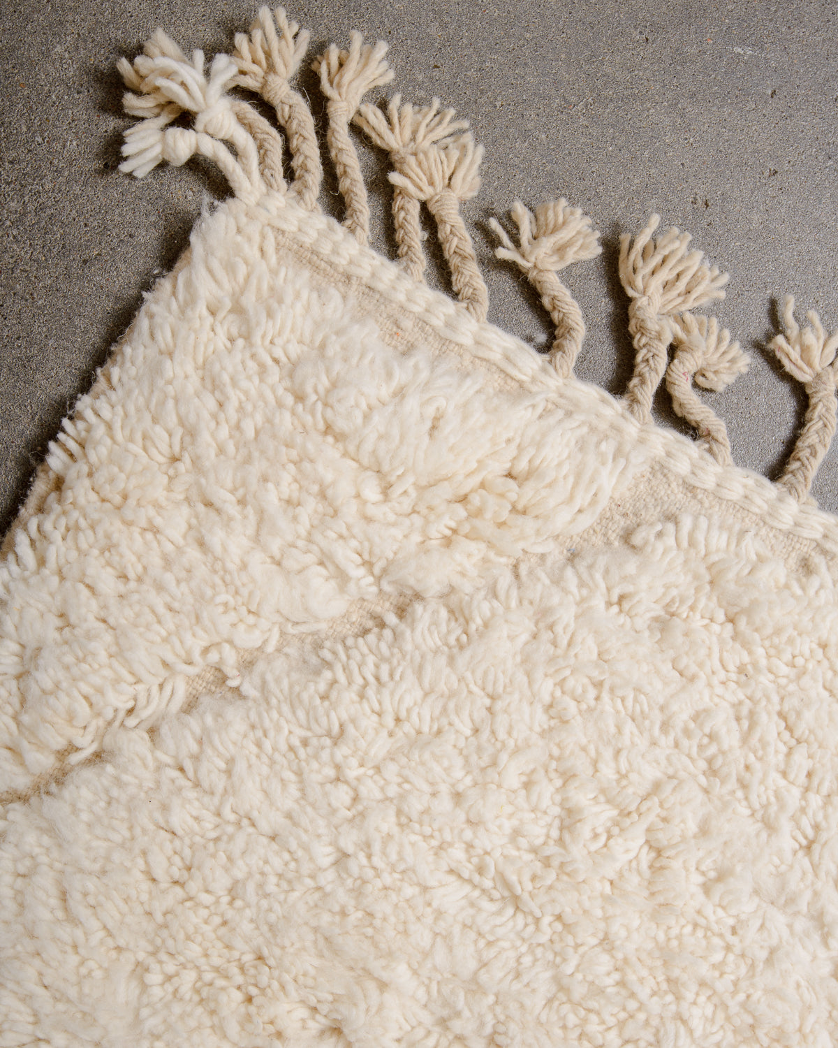 Corner of cream wool rug with tassels on grey cement floor. 