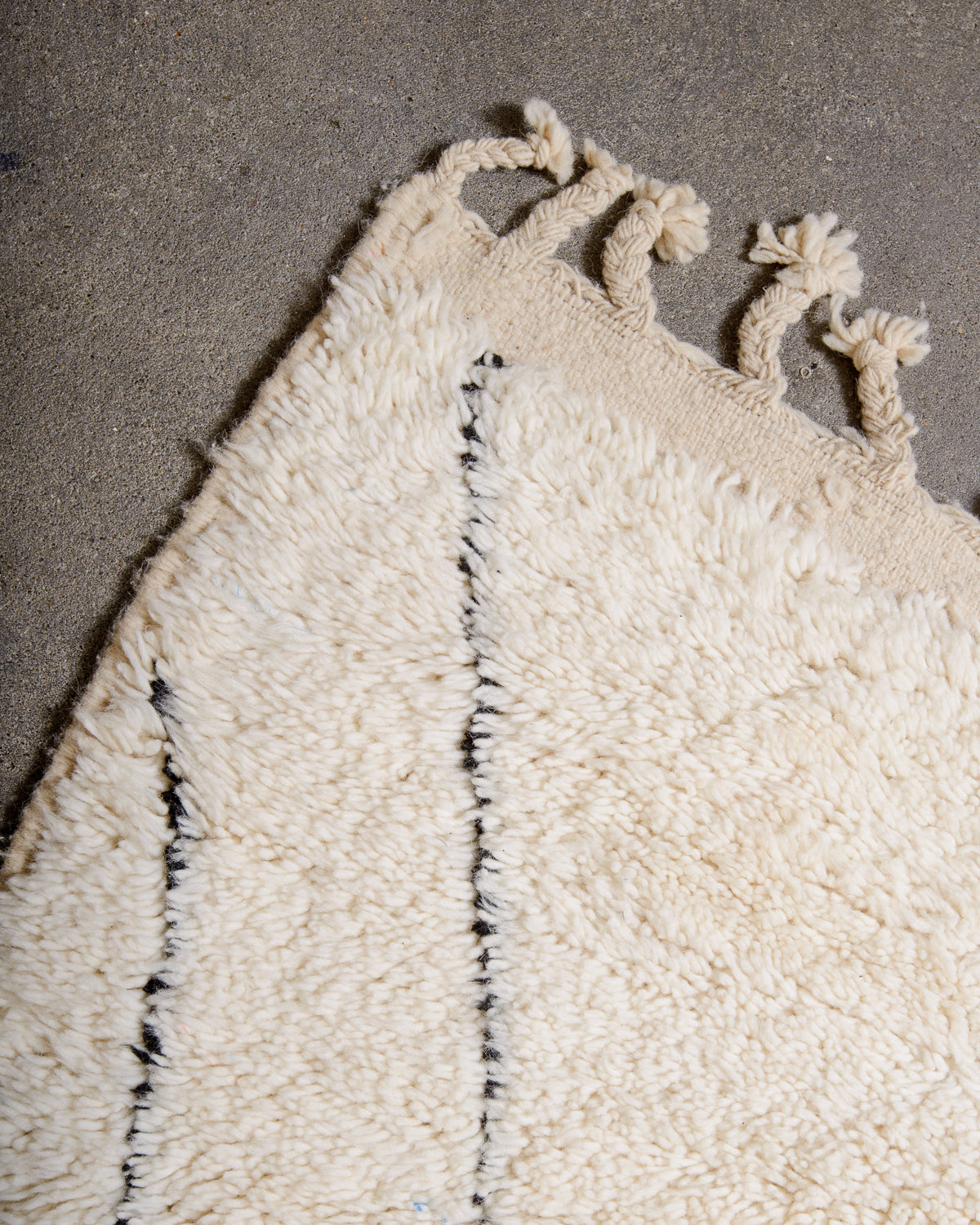 Corner of cream wool rug with tightly woven end and natural wool tassels on grey floor.
