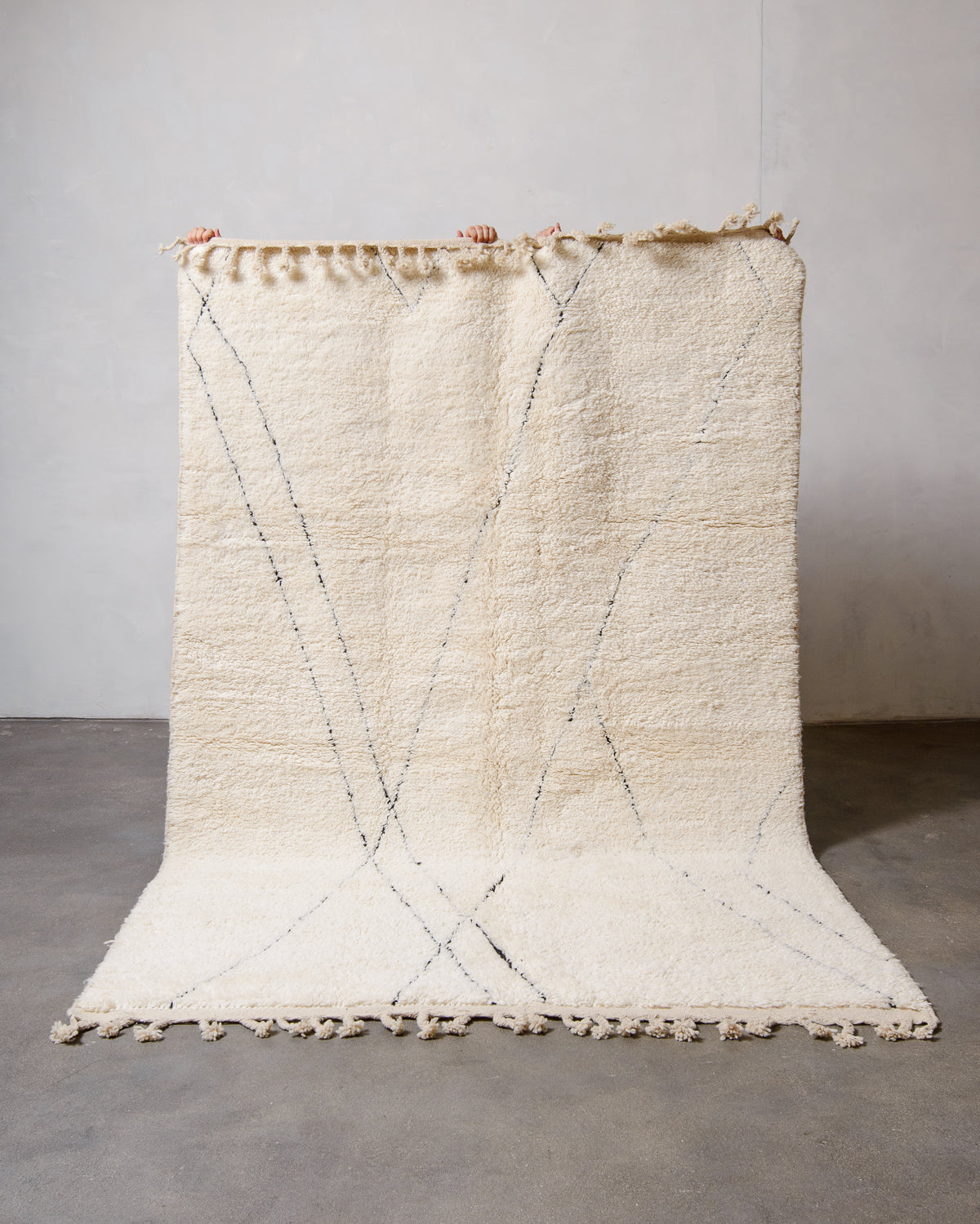 Cream wool rug with black intersecting lines being held up in front of a white wall.