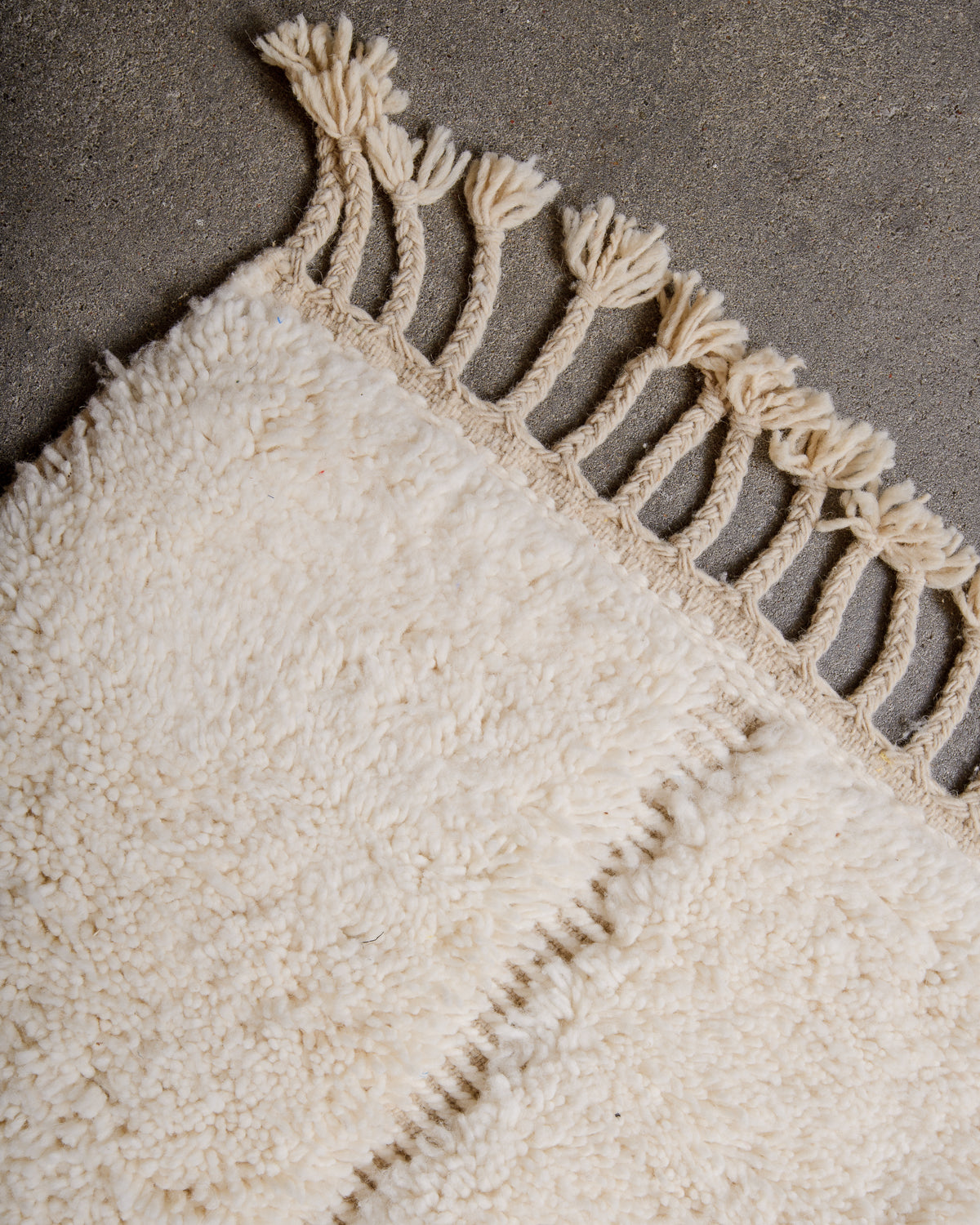 Corner of cream wool rug with tassels on grey cement floor. 