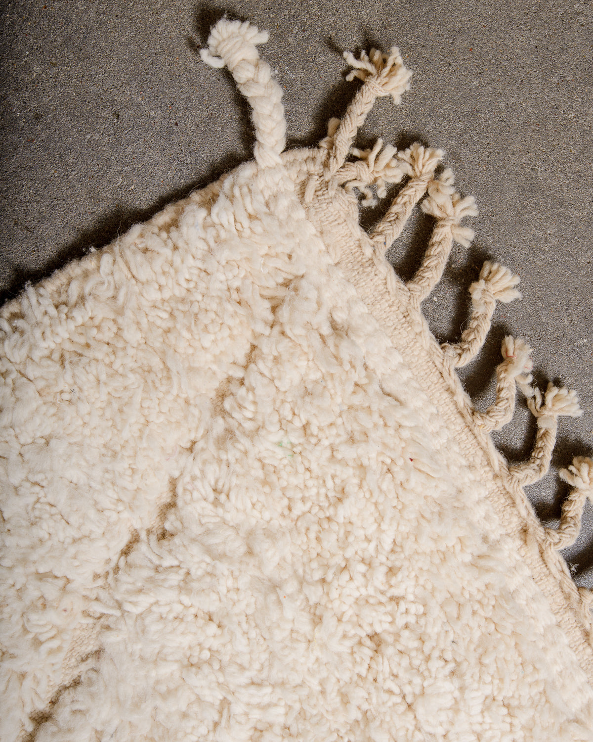 Tasseled edge of Beniourain rug in natural cream with a subtle diamond design, laying on concrete floor.