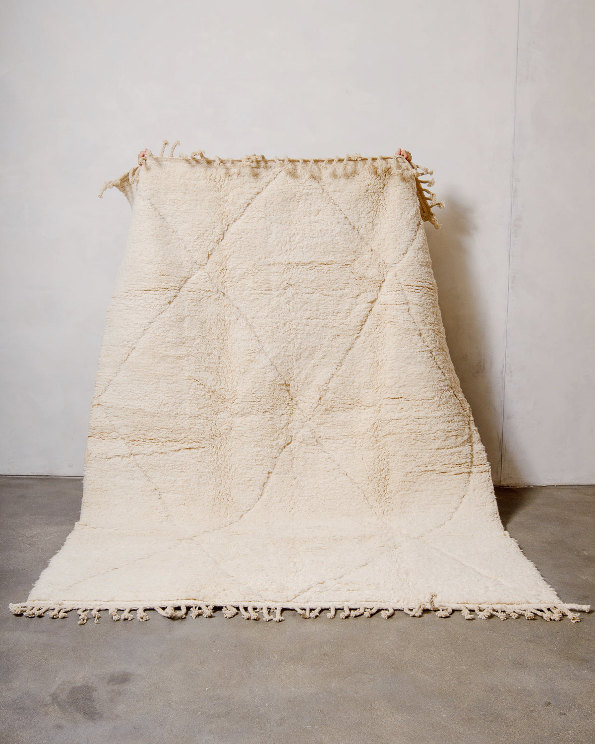 Wool Beniourain rug in natural cream with a subtle diamond design, being held up against a white wall. 