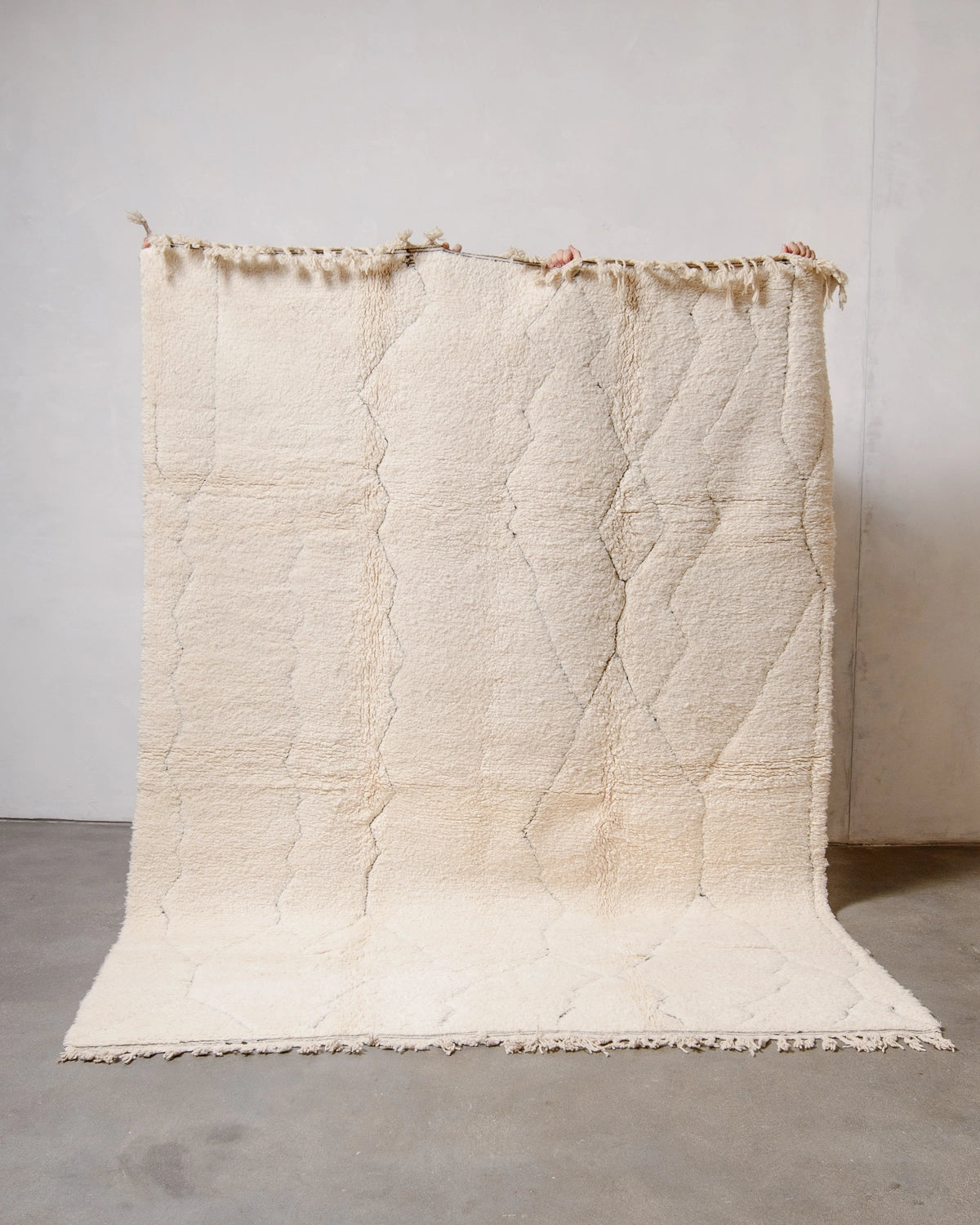 Berberlin Beniourain Rug made of cream wool with a subtle woven design in black, held up against a white studio wall.