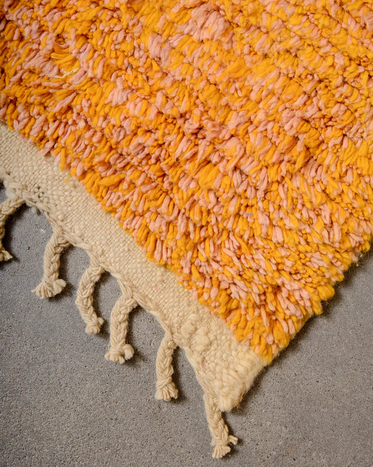 Tasseled edge of a vintage Berber rug made of pink and orange wool laying on a concrete floor.