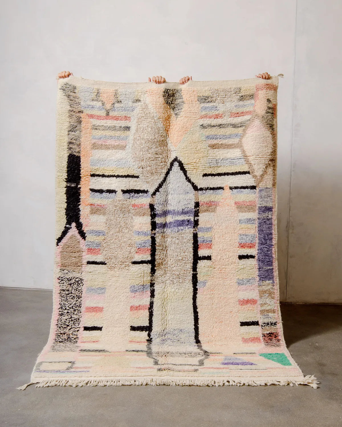 Handcrafted Berber rug from Morocco in shades of soft brown, cream, orange, yellow and purple, being held up against a white wall.