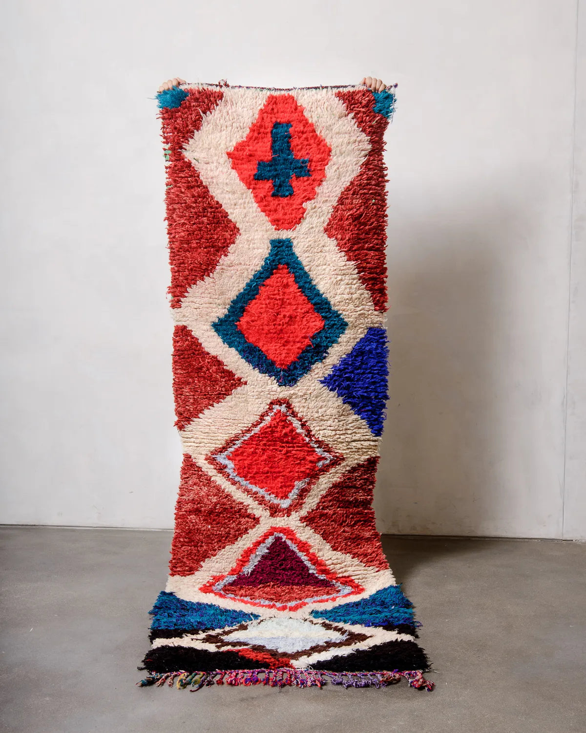 Runner rug with design in predominantly red, cream and blue being held up by two hands in front of a white wall and grey concrete floor. 