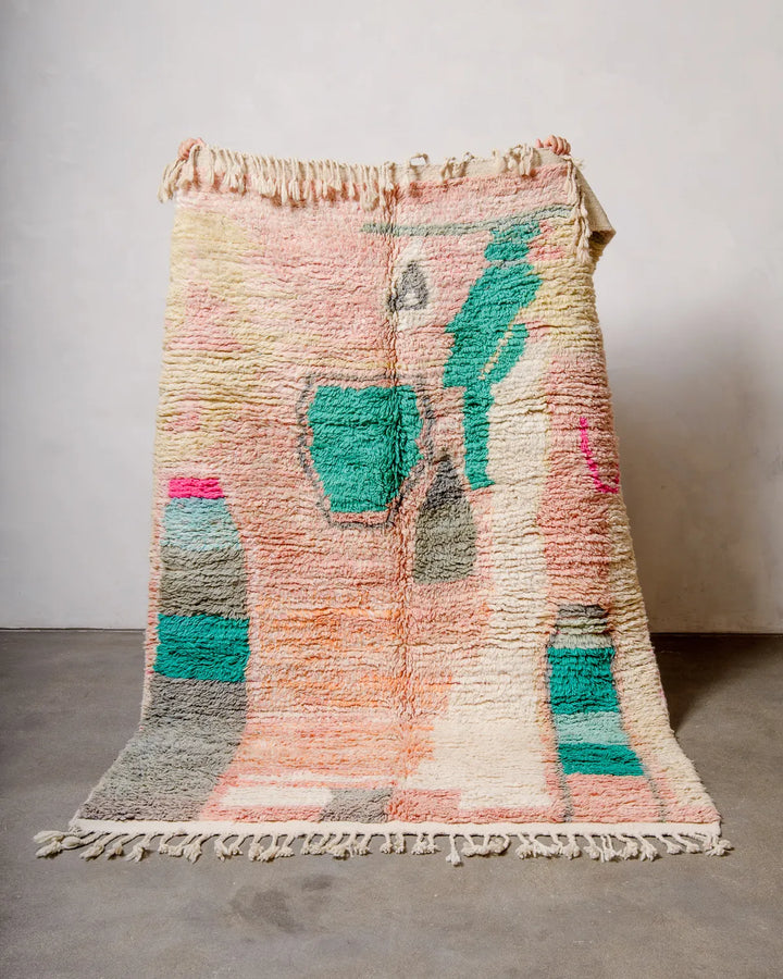 Handcrafted Berber rug from Morocco being held up against white wall. Vintage carpet featuring shades of pink, peach, yellow and turquoise and a fluffy texture.