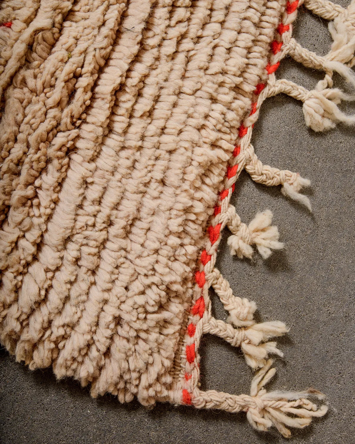 Tasseled edge of vintage wool rug in shades of sand and bright tomato red, laying on cement floor.