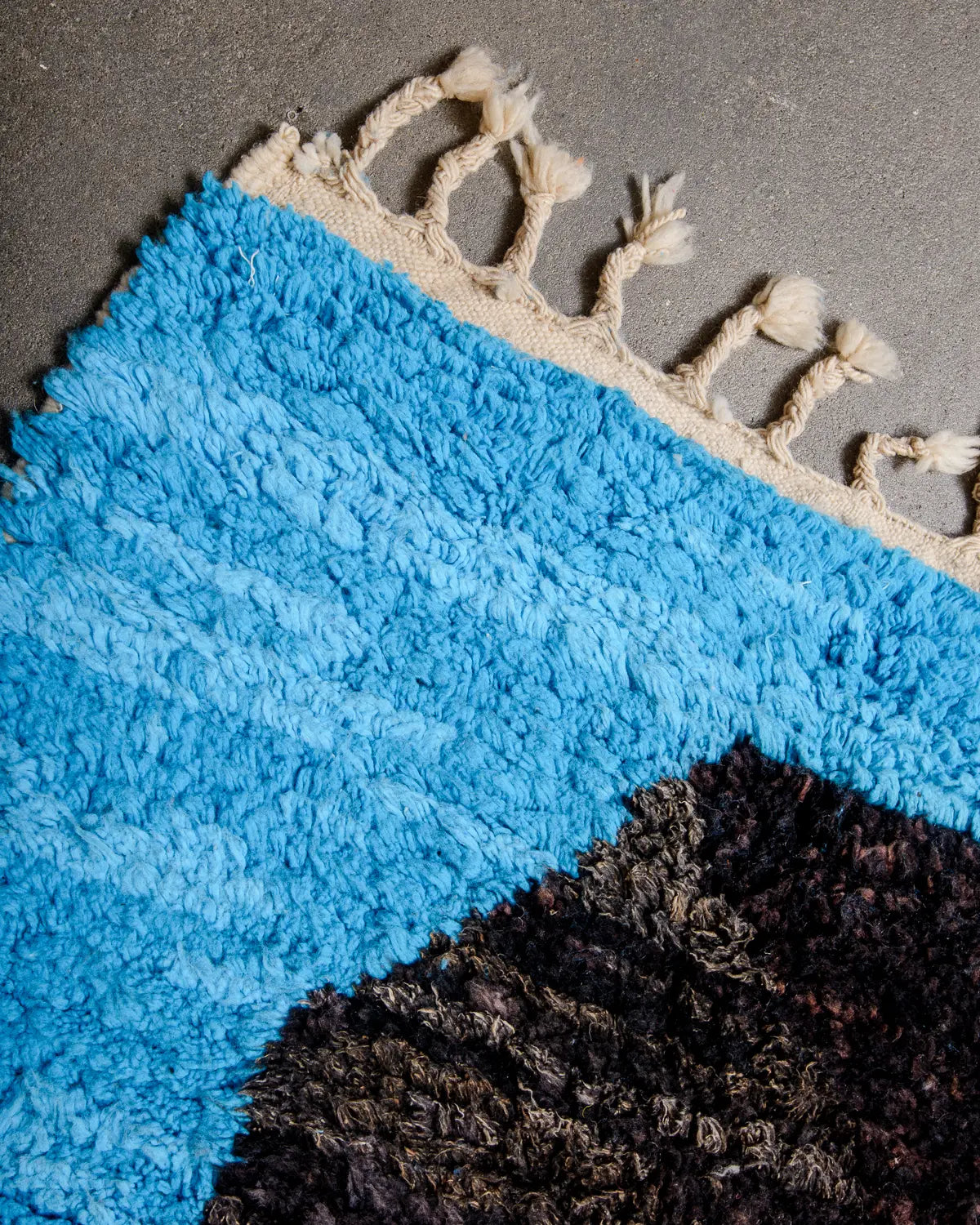 Tasseled edge of vintage Berber rug in shades of black and blue with natural wool edge and tassels.