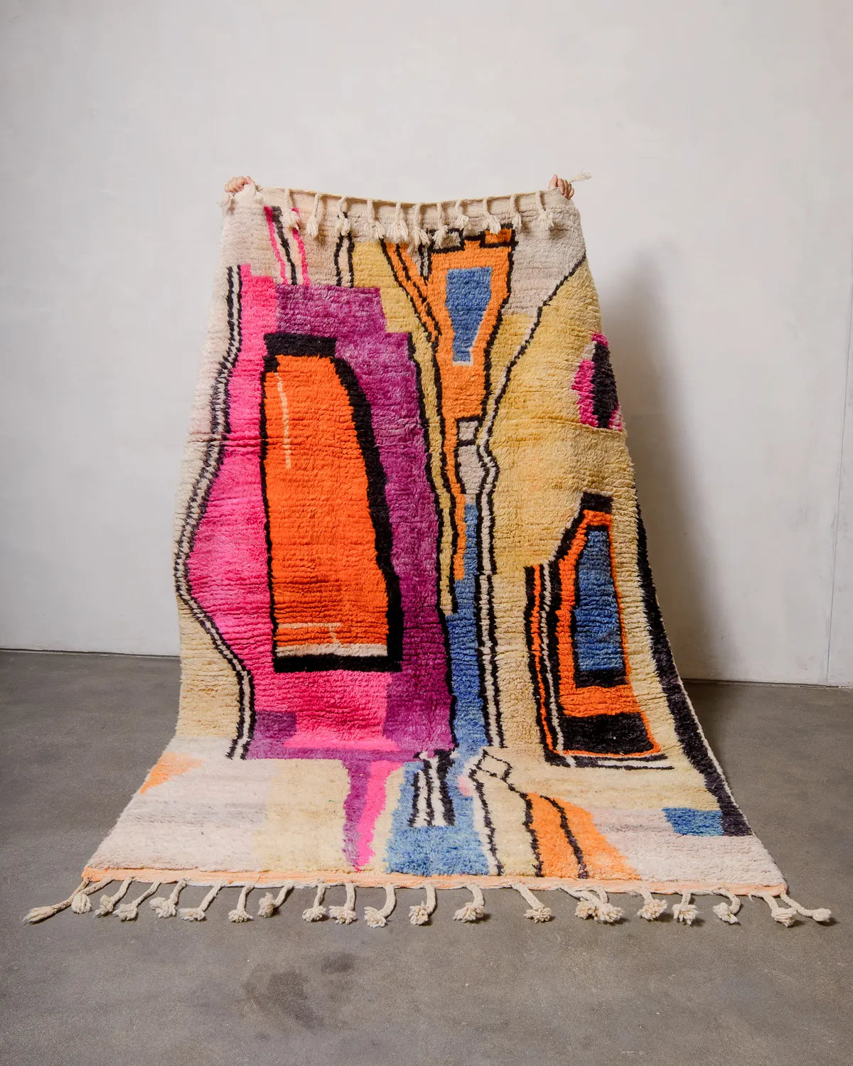 Handwoven Berberlin Berber rug from Morocco being held up against a white wall. Vintage carpet featuring a modern design in shades of pink, yellow, orange, blue and black.