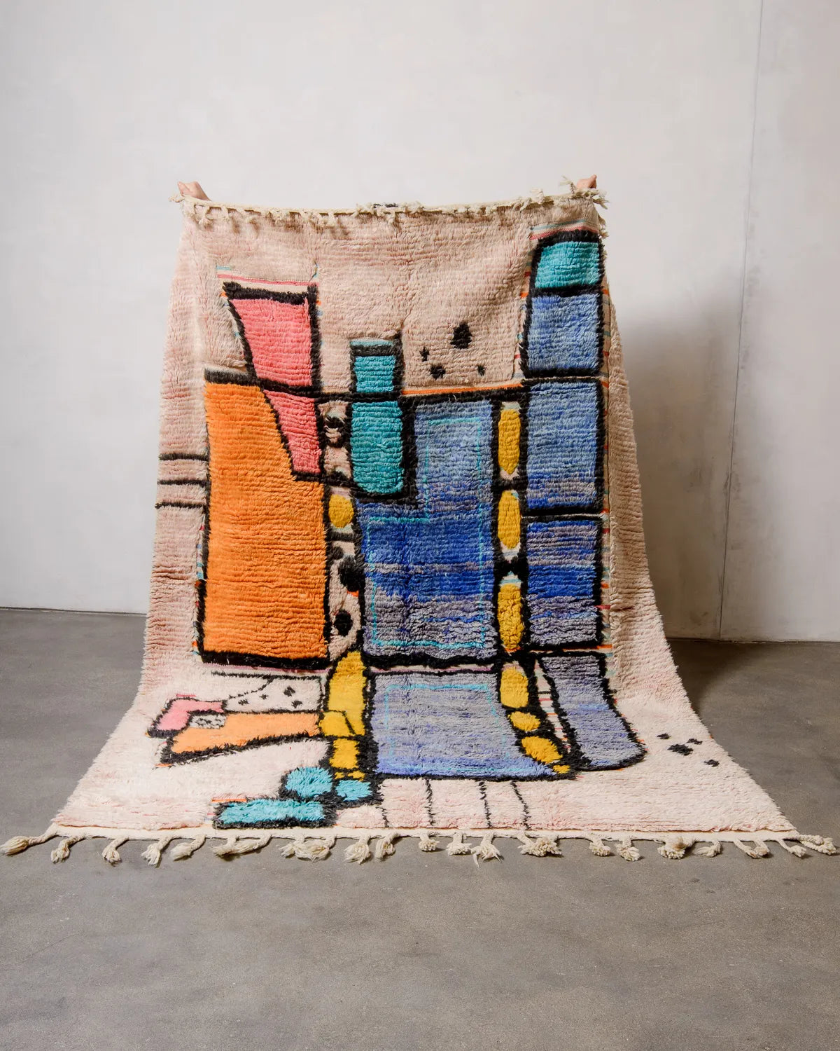 Handwoven Berberlin Boujad rug being held up against a white wall. The Moroccan rug features fluffy wool in shades of blue, turquoise, orange, yellow, pink and black, with flat-woven sections in stripes. 