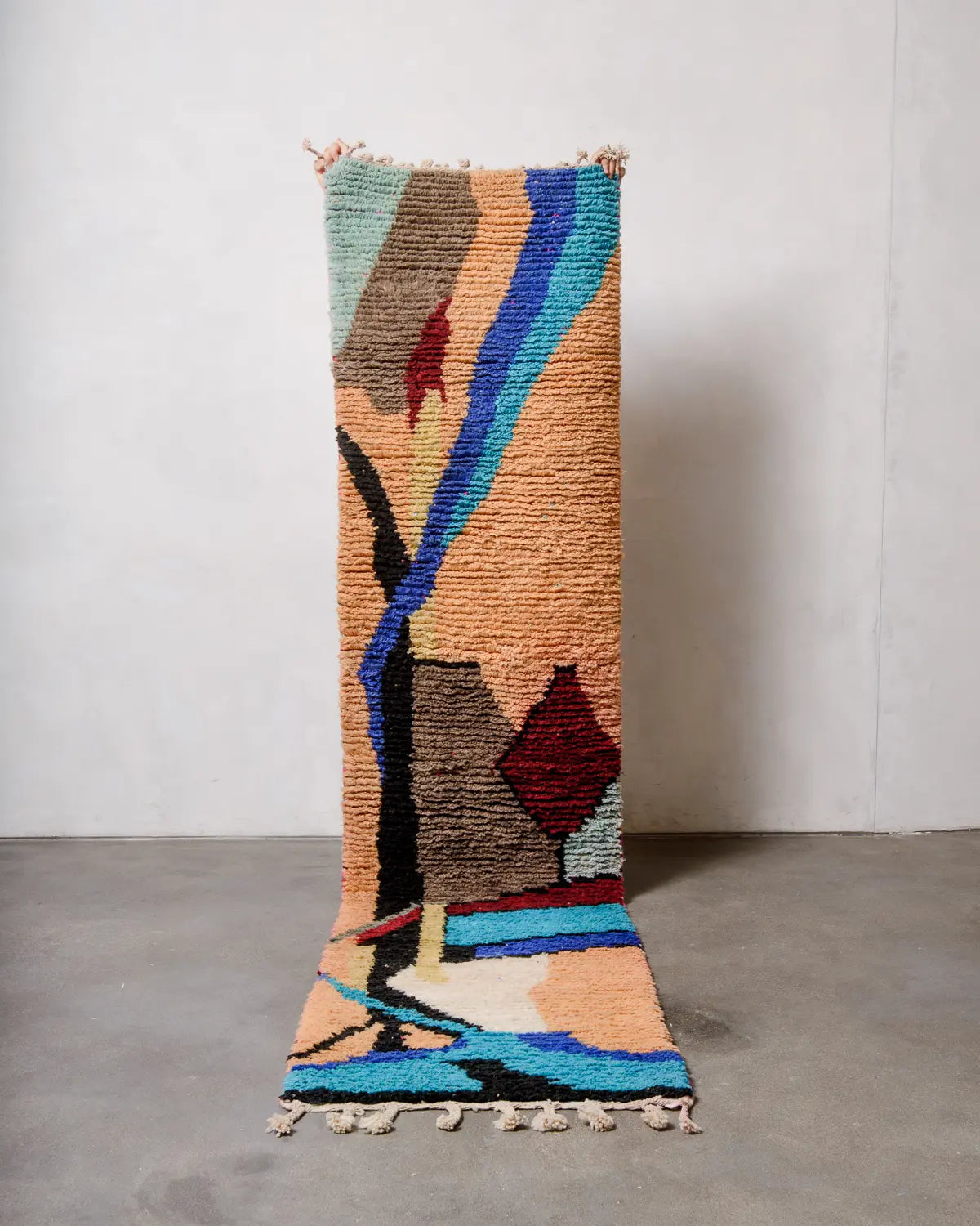 Wool Berberlin runner rug being held up in front of white studio wall and grey concrete floor.