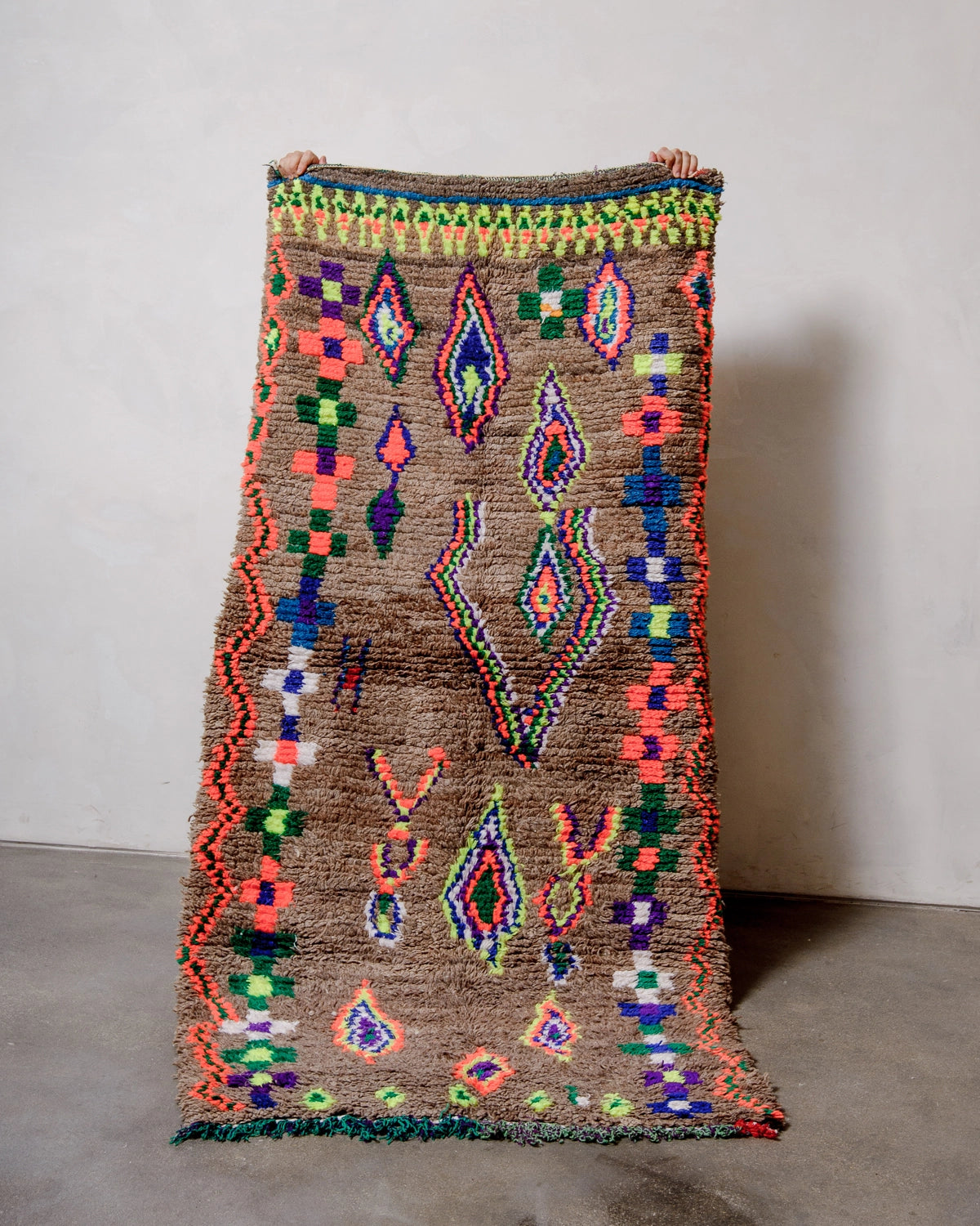 Vintage Berber Rug made of brown wool with design details in neon, purple, blue, green cotton,  held up against a white wall. 