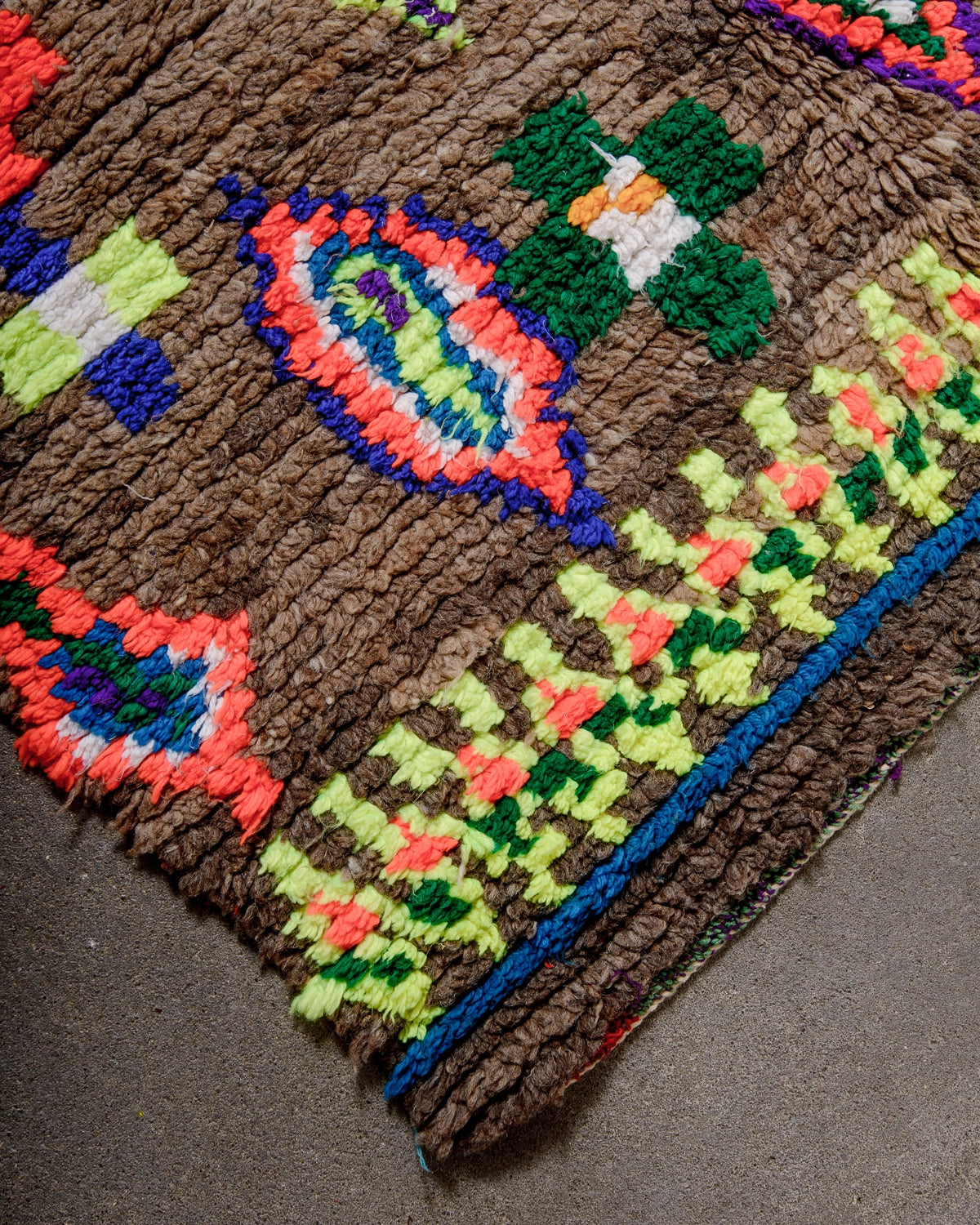 Detail of Vintage Berber Rug made of brown wool with design details in neon, purple, blue, green cotton laying on concrete.