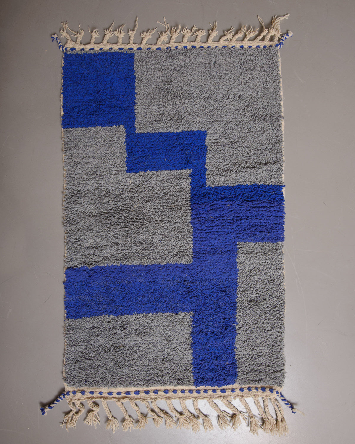 Grey and blue rug with cream tassels on grey floor.