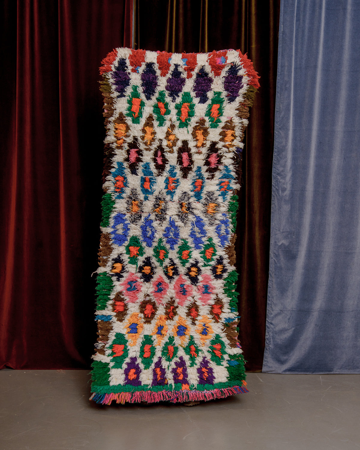 Vintage Berberlin boucherouite runner rug being held up in front of a velvet curtain. 