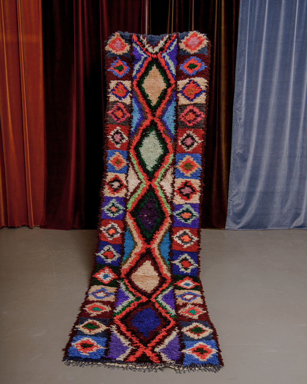 Vintage Berberlin boucherouite runner rug being held up in front of a velvet curtain. 