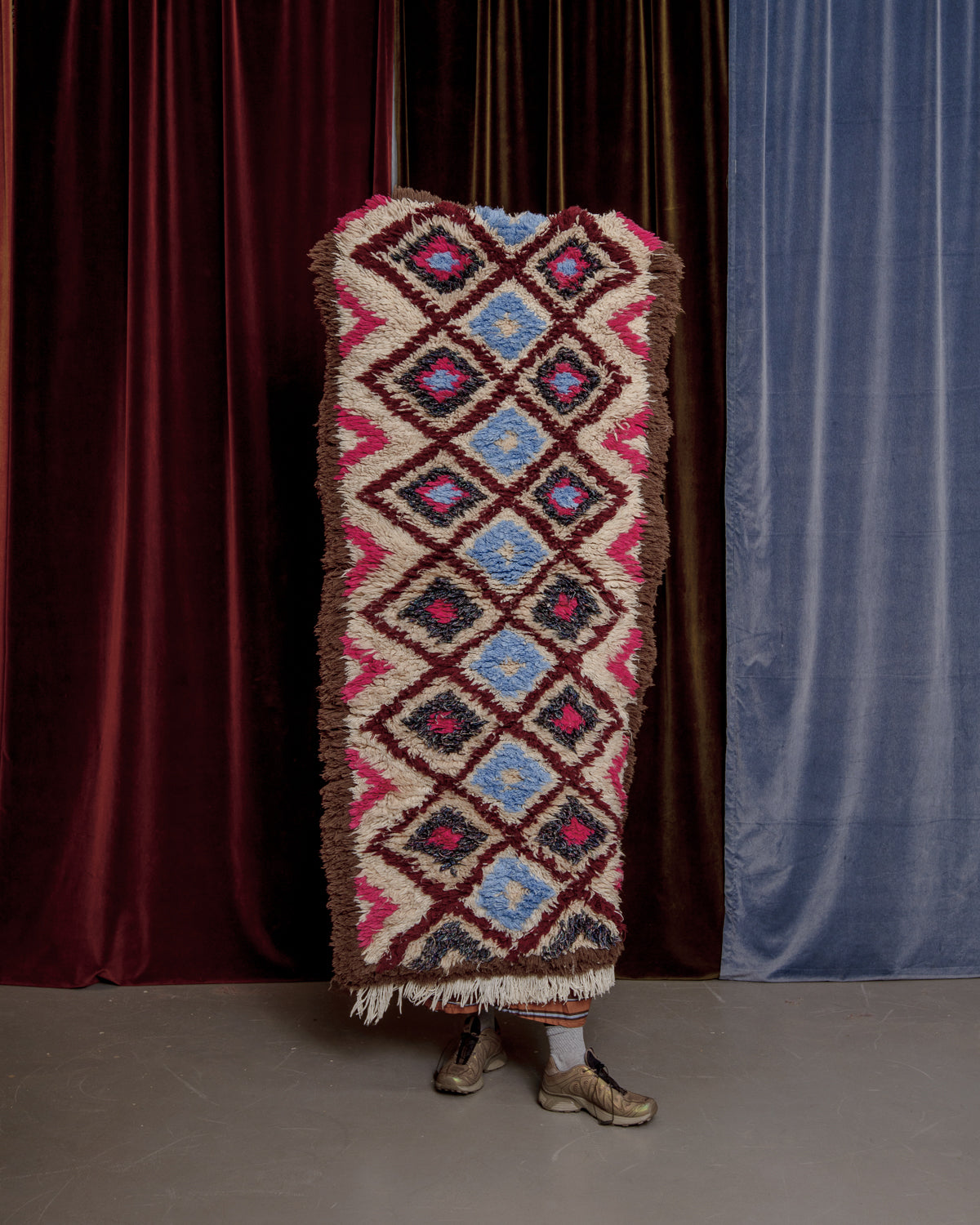 Vintage Berberlin boucherouite runner rug being held up in front of a velvet curtain. 