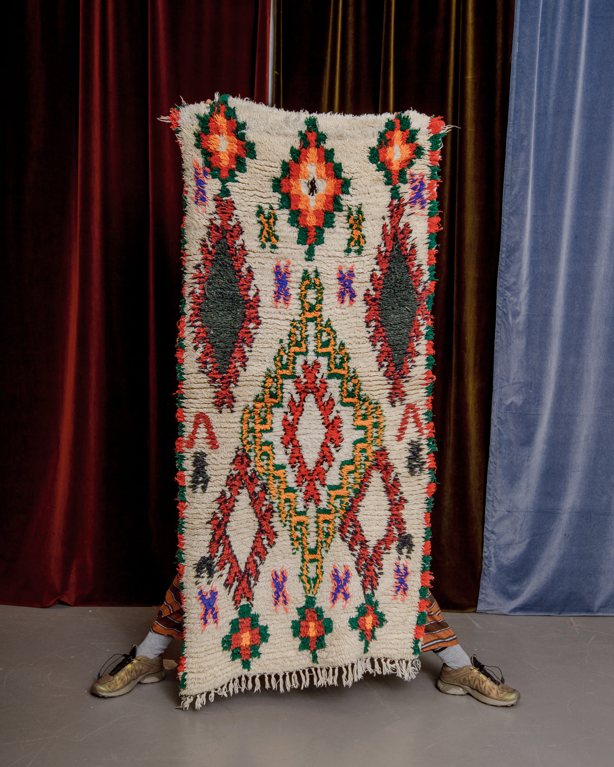 Vintage Berberlin boucherouite runner rug being held up in front of a velvet curtains.