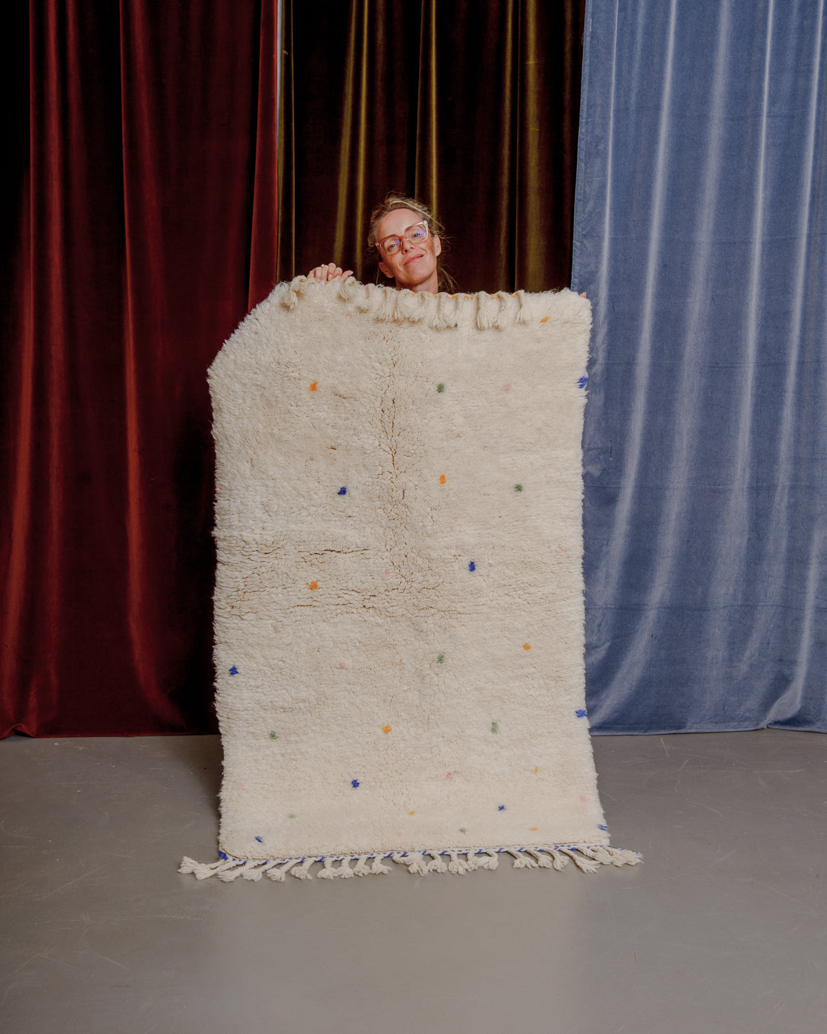 Cream wool rug with coloured dots being held by a woman in large glasses in front of velvet curtains.