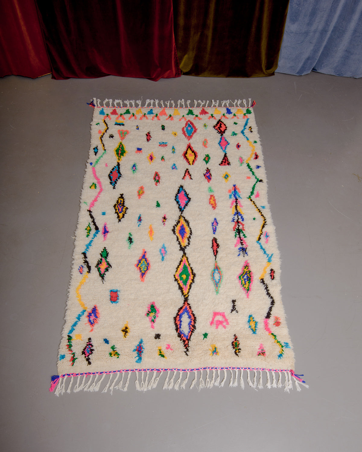 Azilal rug with colourful, abstract design laid flat on grey floor in front of a wall of velvet curtains.