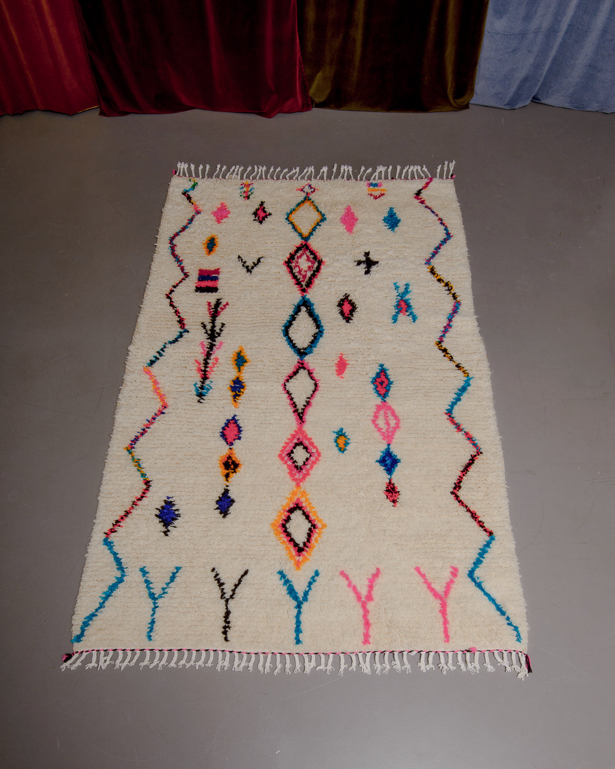 Azilal rug with colourful, abstract design laid flat on grey floor in front of a wall of velvet curtains.