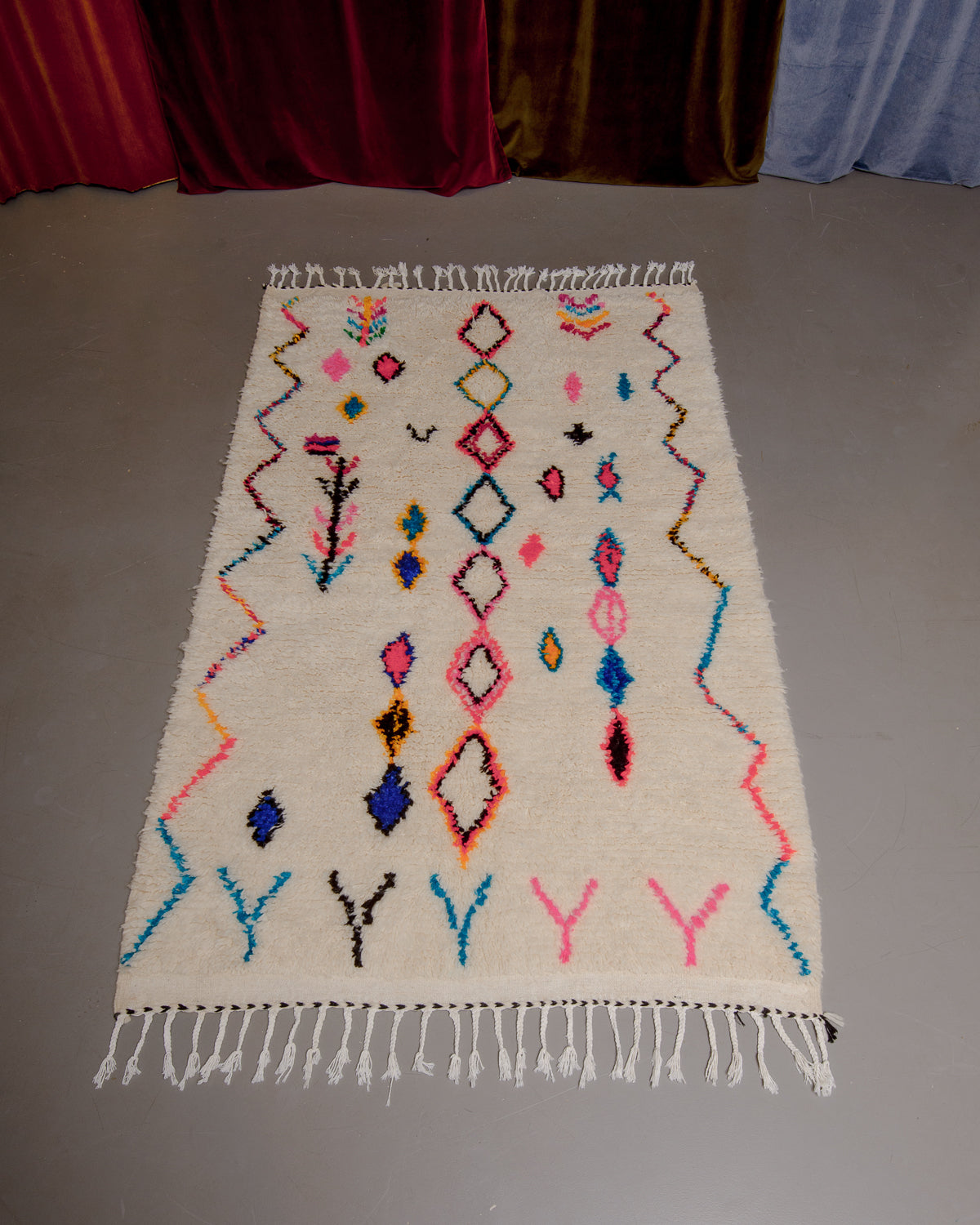 Azilal rug with colourful, abstract design laid flat on grey floor in front of a wall of velvet curtains.