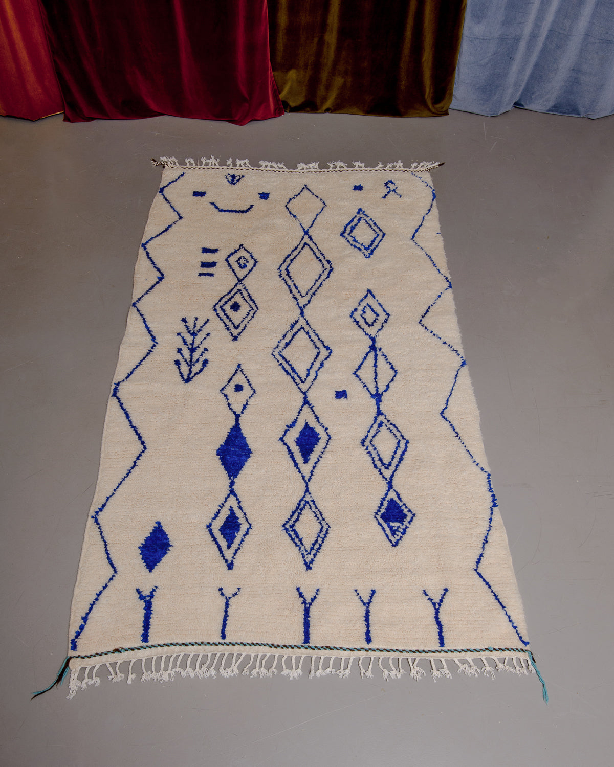 Azilal rug with abstract design in cream and blue, laid flat on grey floor in front of a wall of velvet curtains.