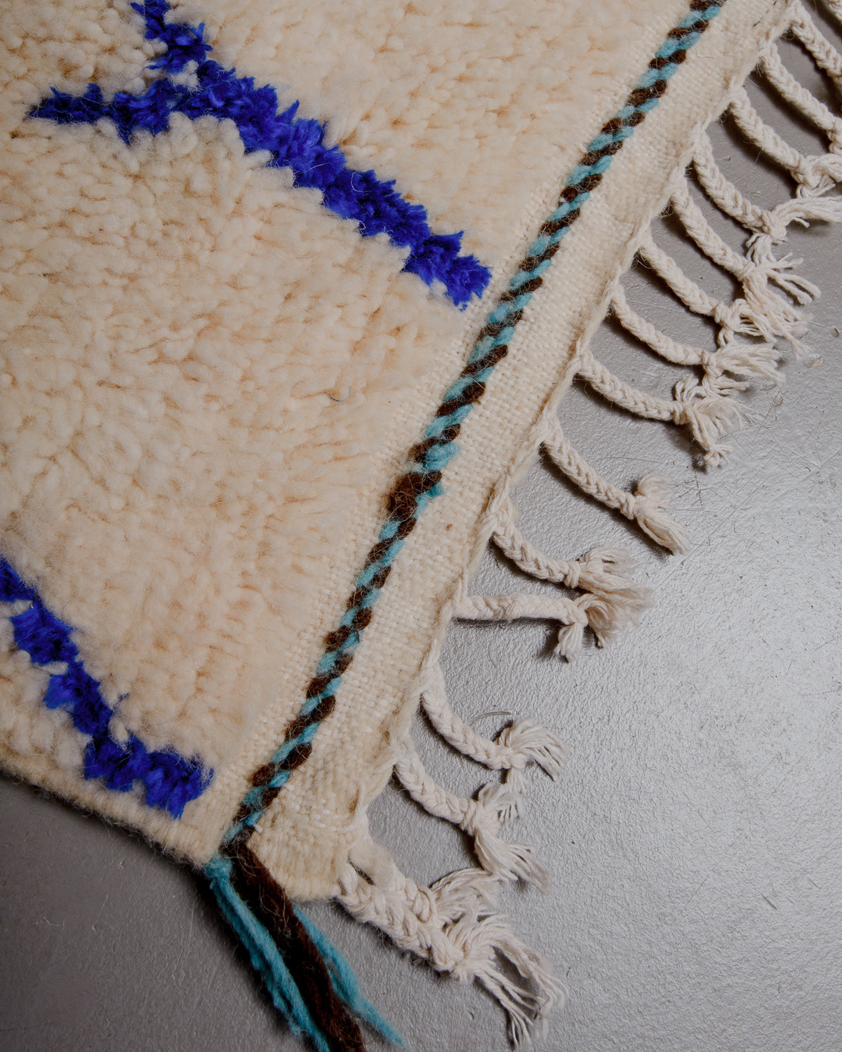 Corner of azilal rug with white cotton tassels and turquoise and brown embroidery along edge.