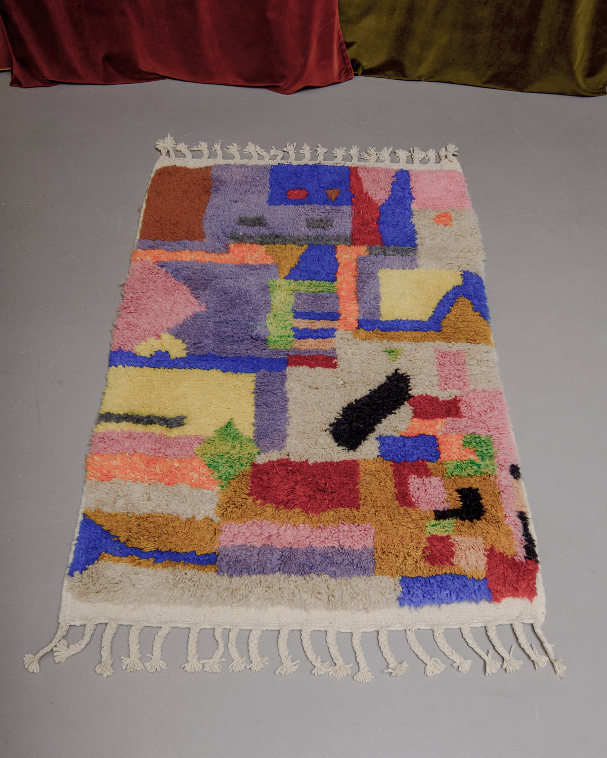 Berberlin Beniourain rug with colourful geometric pattern on grey floor in front of velvet curtains. 