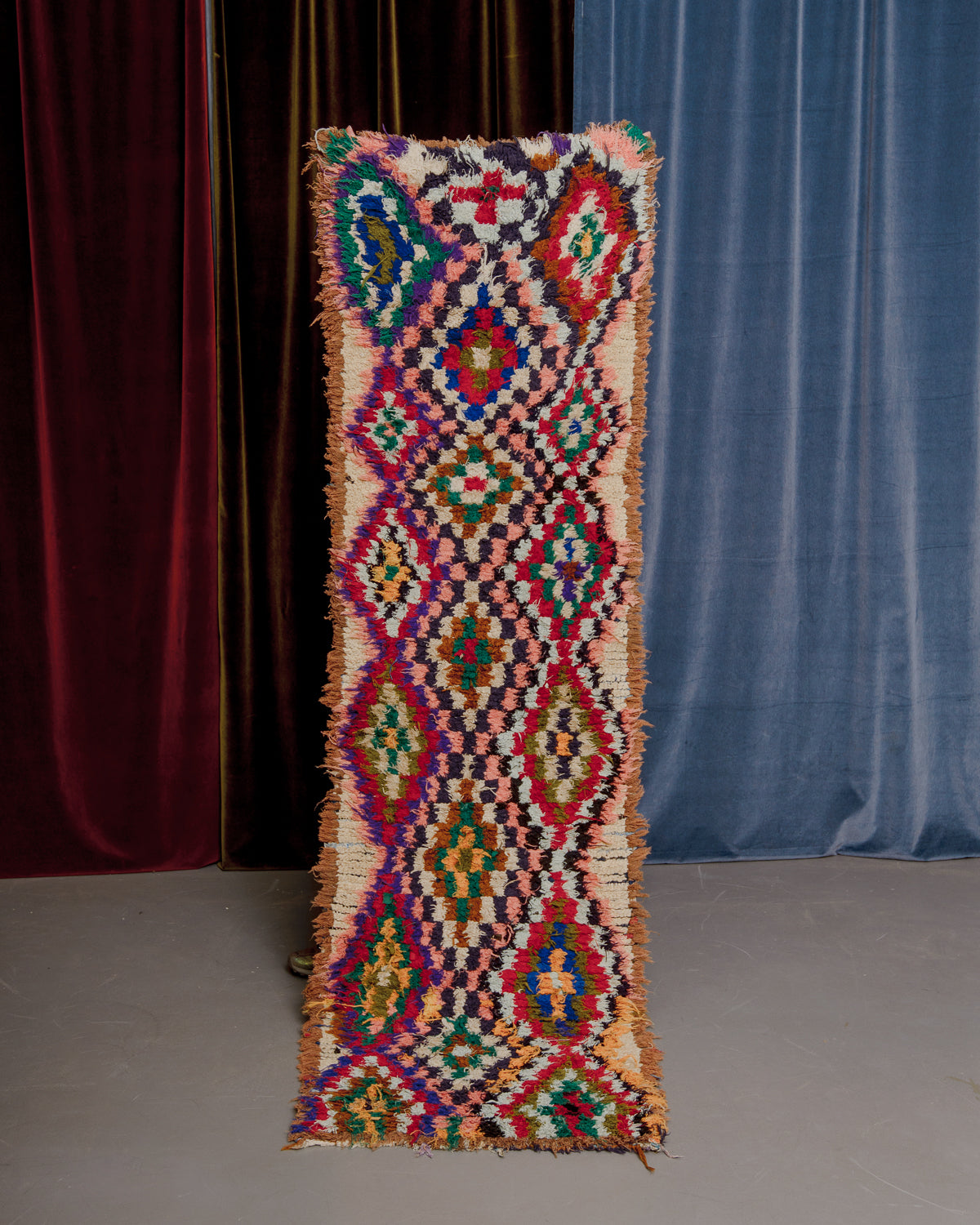 Vintage Berberlin boucherouite runner rug being held up in front of velvet curtains. 