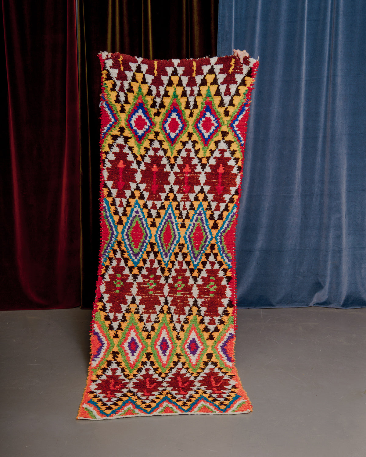 Vintage Berberlin boucherouite runner rug being held up in front of velvet curtains. 
