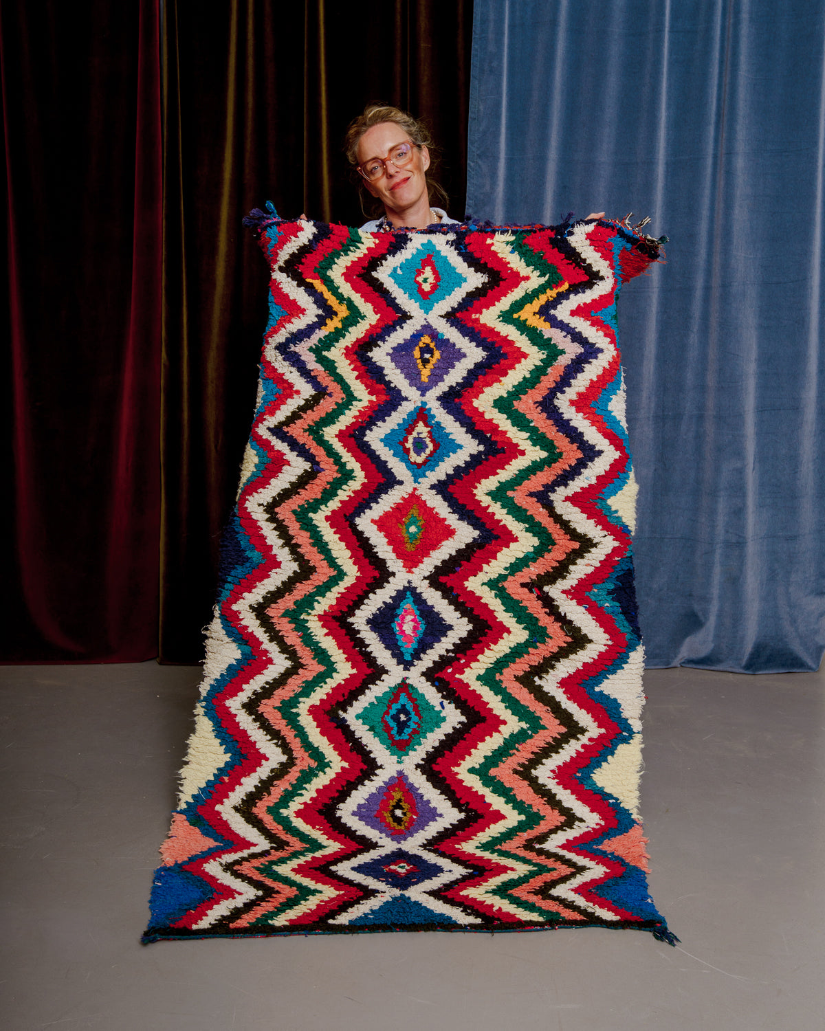 Vintage Berberlin boucherouite runner rug being held up in front of velvet curtains. 