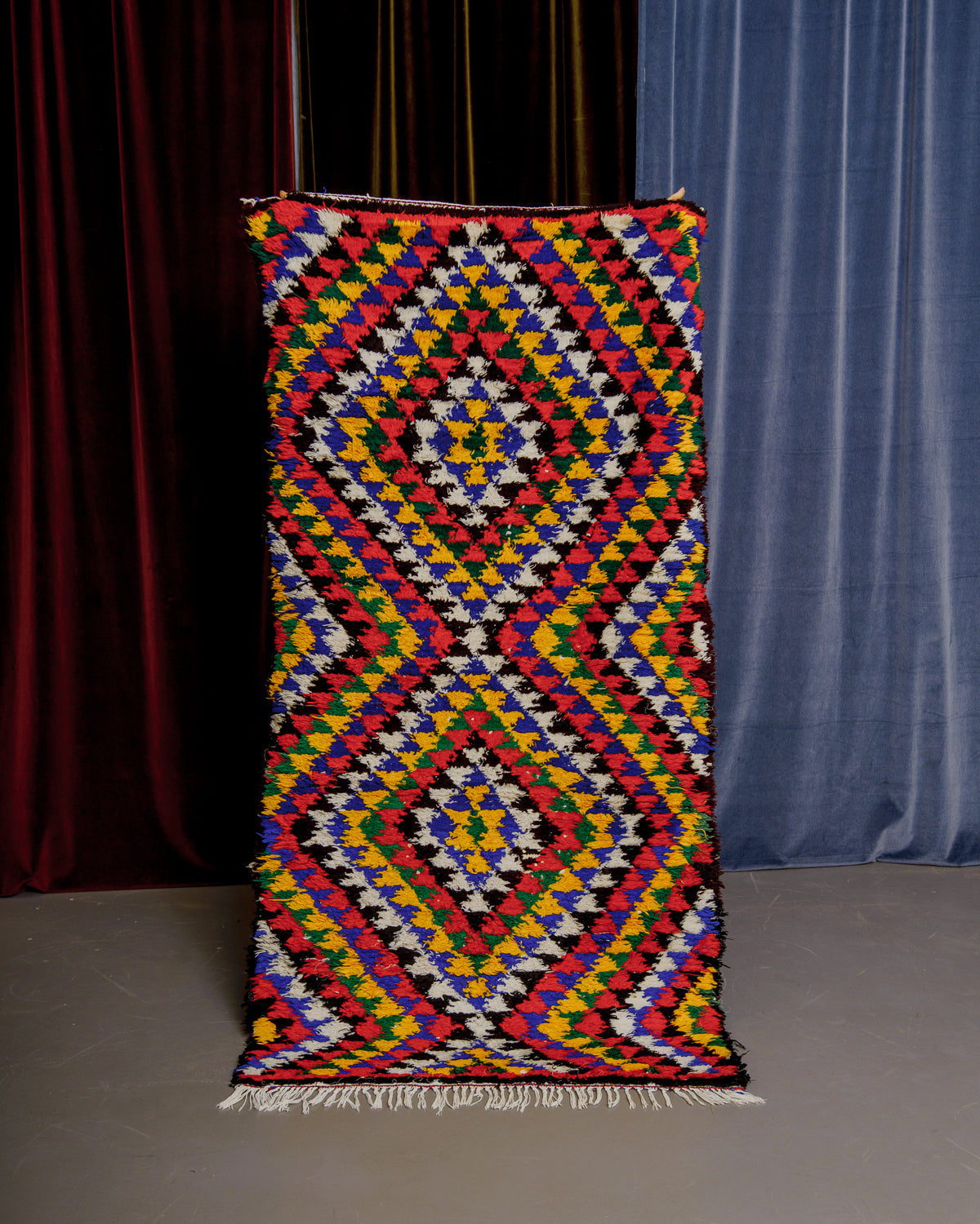 Vintage Berberlin boucherouite runner rug being held up in front of velvet curtains. 