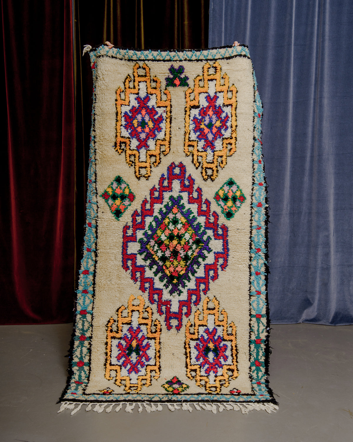Vintage Berberlin boucherouite runner rug being held up in front of a velvet curtains.