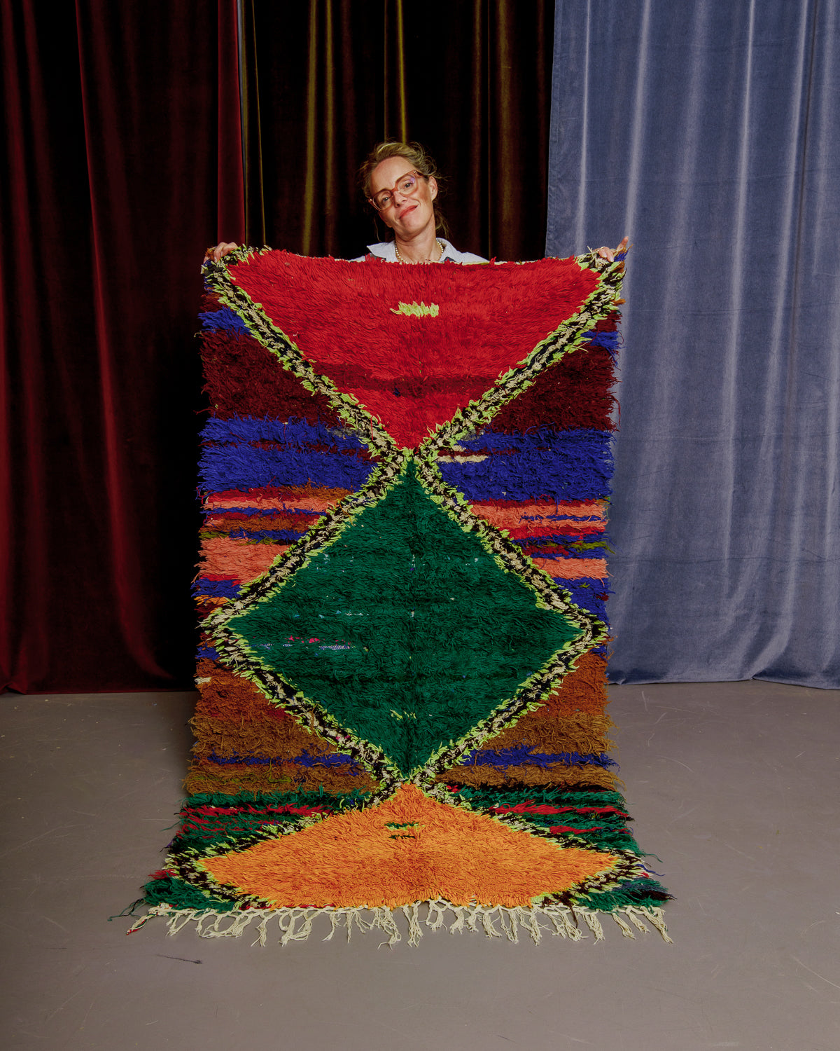Vintage Berberlin boucherouite runner rug being held up by woman in glasses in front of velvet curtains. 