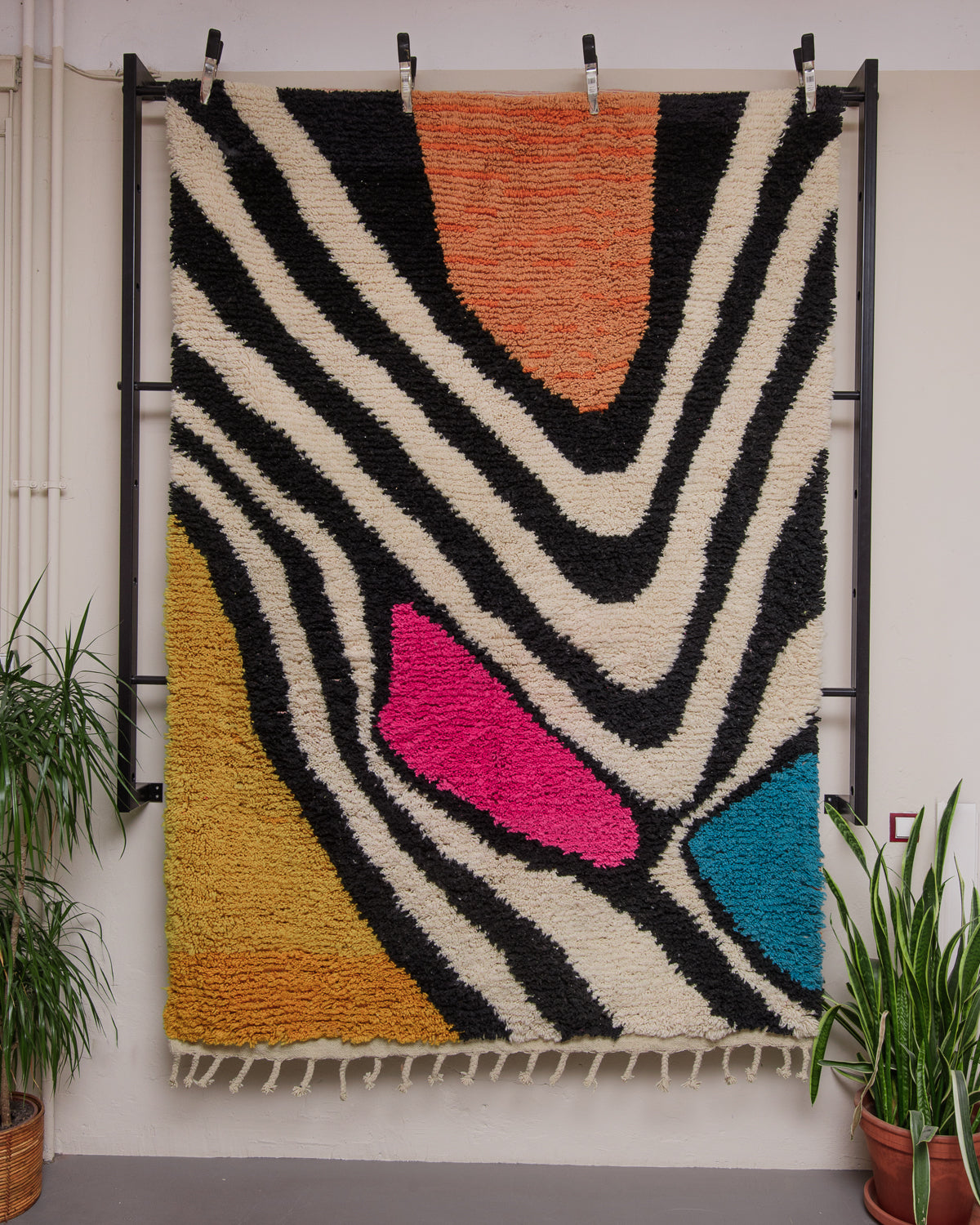 Berberlin Boujaad rug in pink, orange,  yellow, black, white and turquoise wool hung vertically on a frame against a white wall with some green indoor plants.