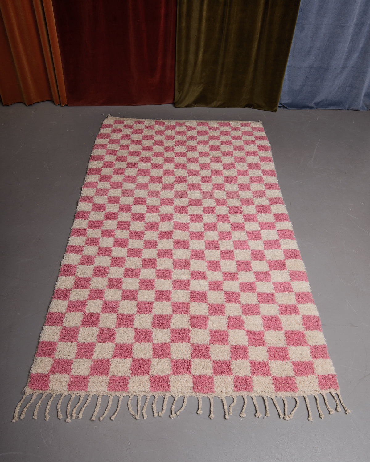 Berberlin Boujaad rug in pink and cream checkered wool laid out on grey floor in front of velvet curtains.