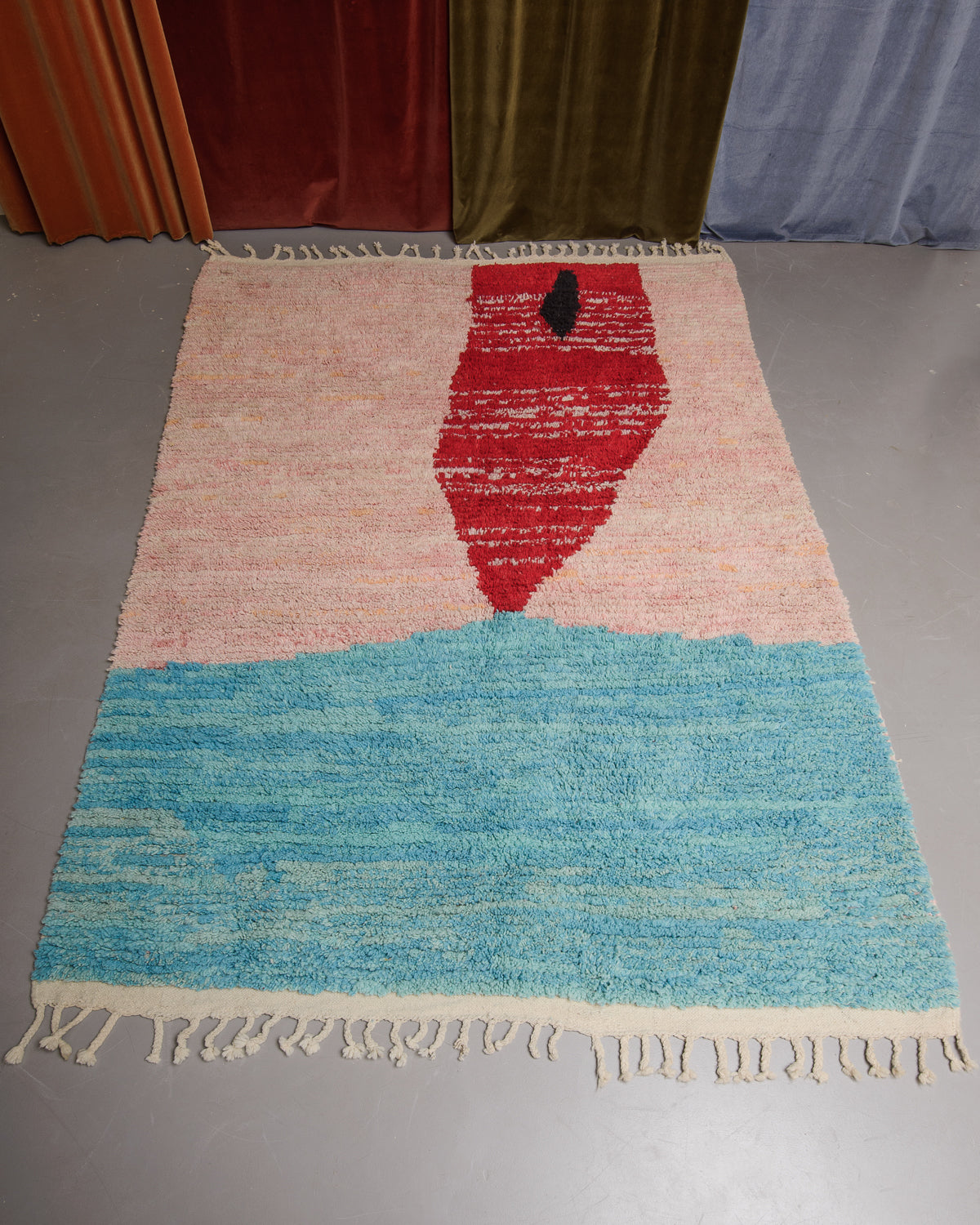 Berberlin Boujaad rug in pink, red and turquoise wool on grey floor in front of velvet curtains.